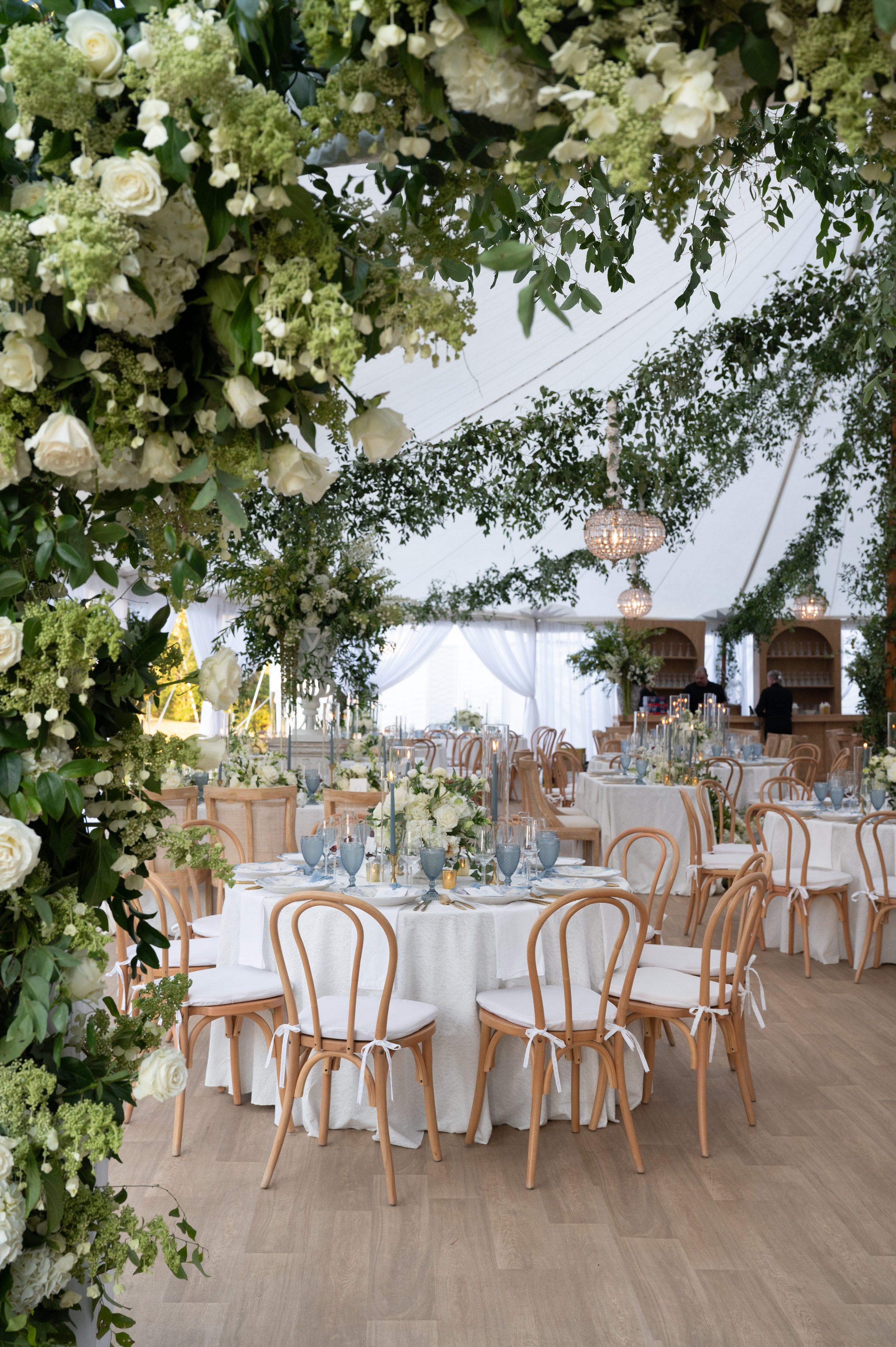 Elegant indoor wedding reception decorated with white flowers, greenery, and chandeliers, featuring round tables with white tablecloths, white floral centerpieces, gold accents, and wooden chairs with white cushions.