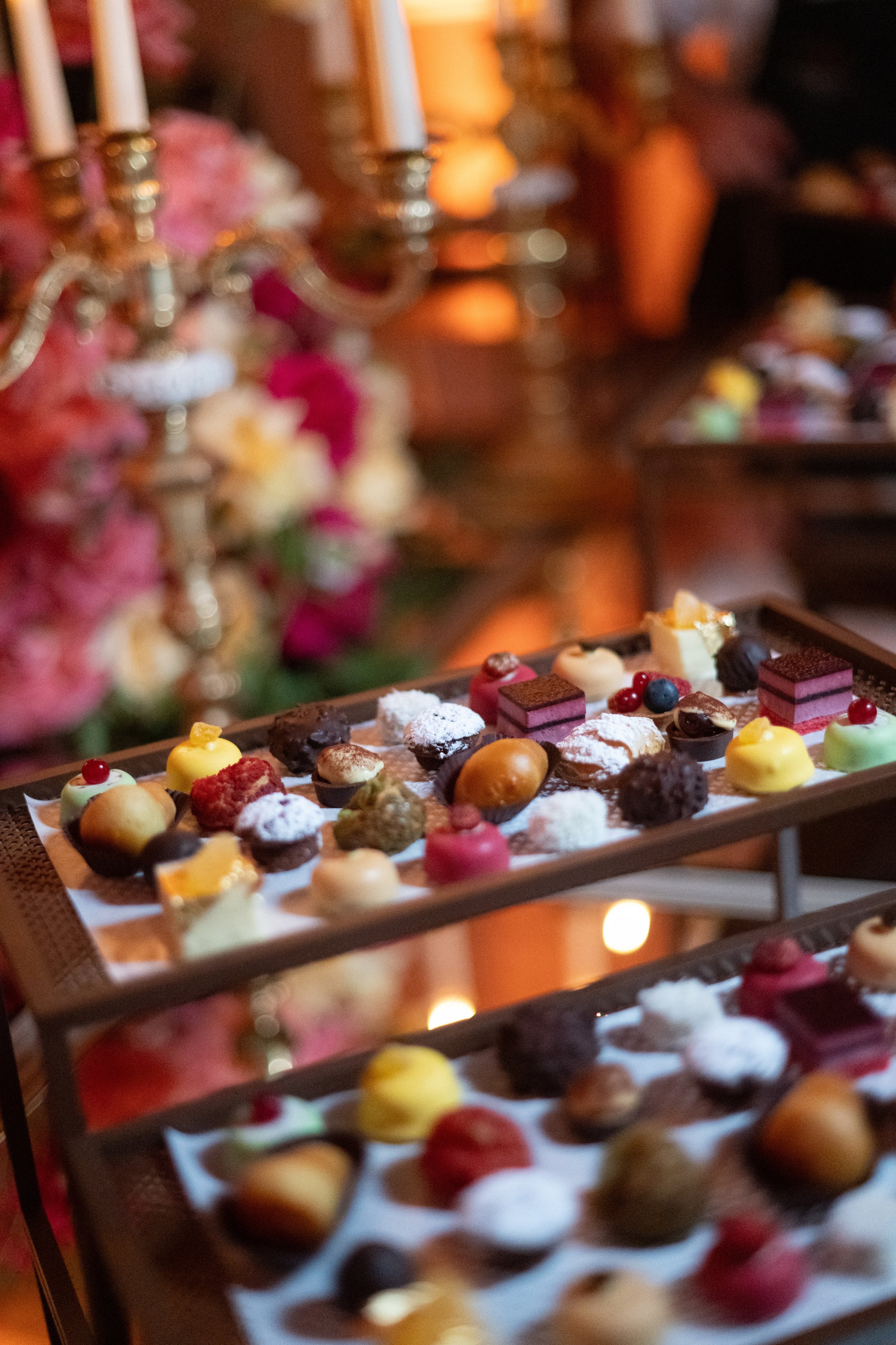 Close-up of a tray filled with assorted small desserts, including cakes, pastries, and chocolates, with a blurred background of candles and flower arrangements.