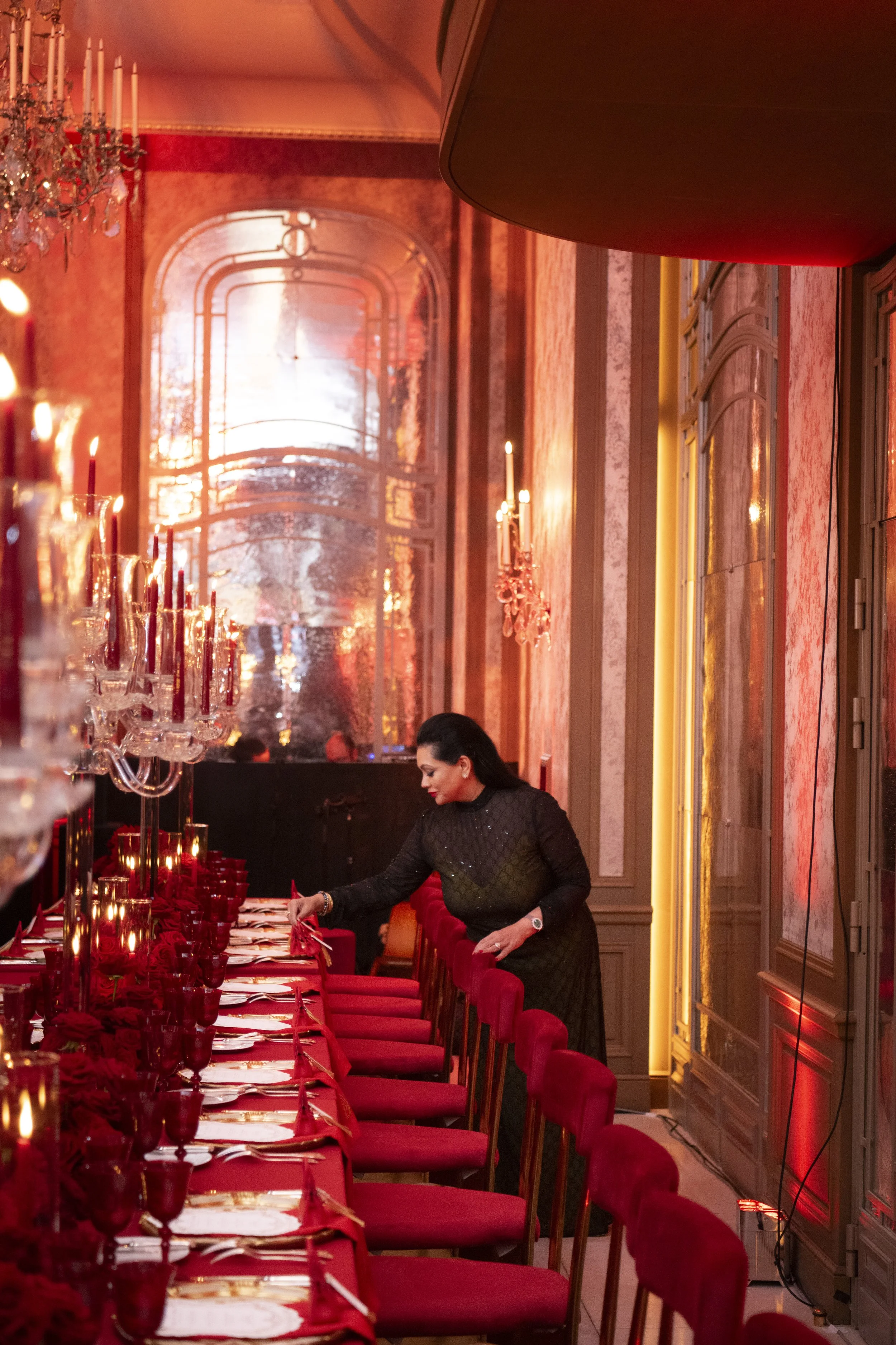 A woman in a black dress setting a table with red tablecloths, red napkins, and red glassware in an elegant, dimly lit room decorated for a formal event.