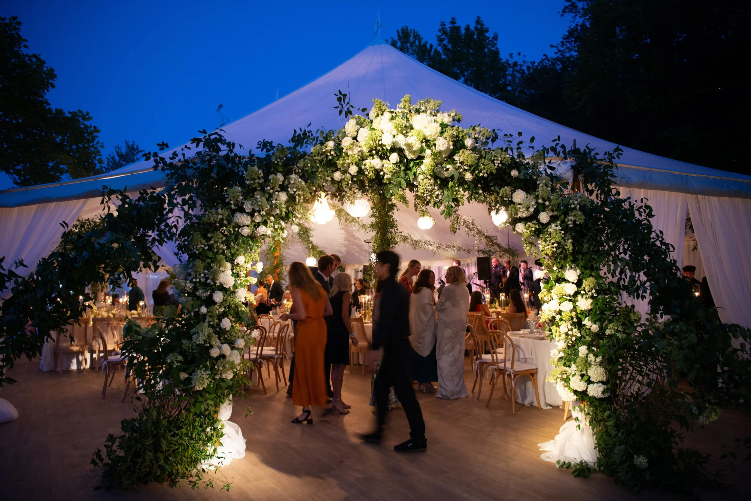 Wedding reception under a large white tent decorated with a floral arch of white flowers. Guests are socializing and mingling inside the tent during evening time.