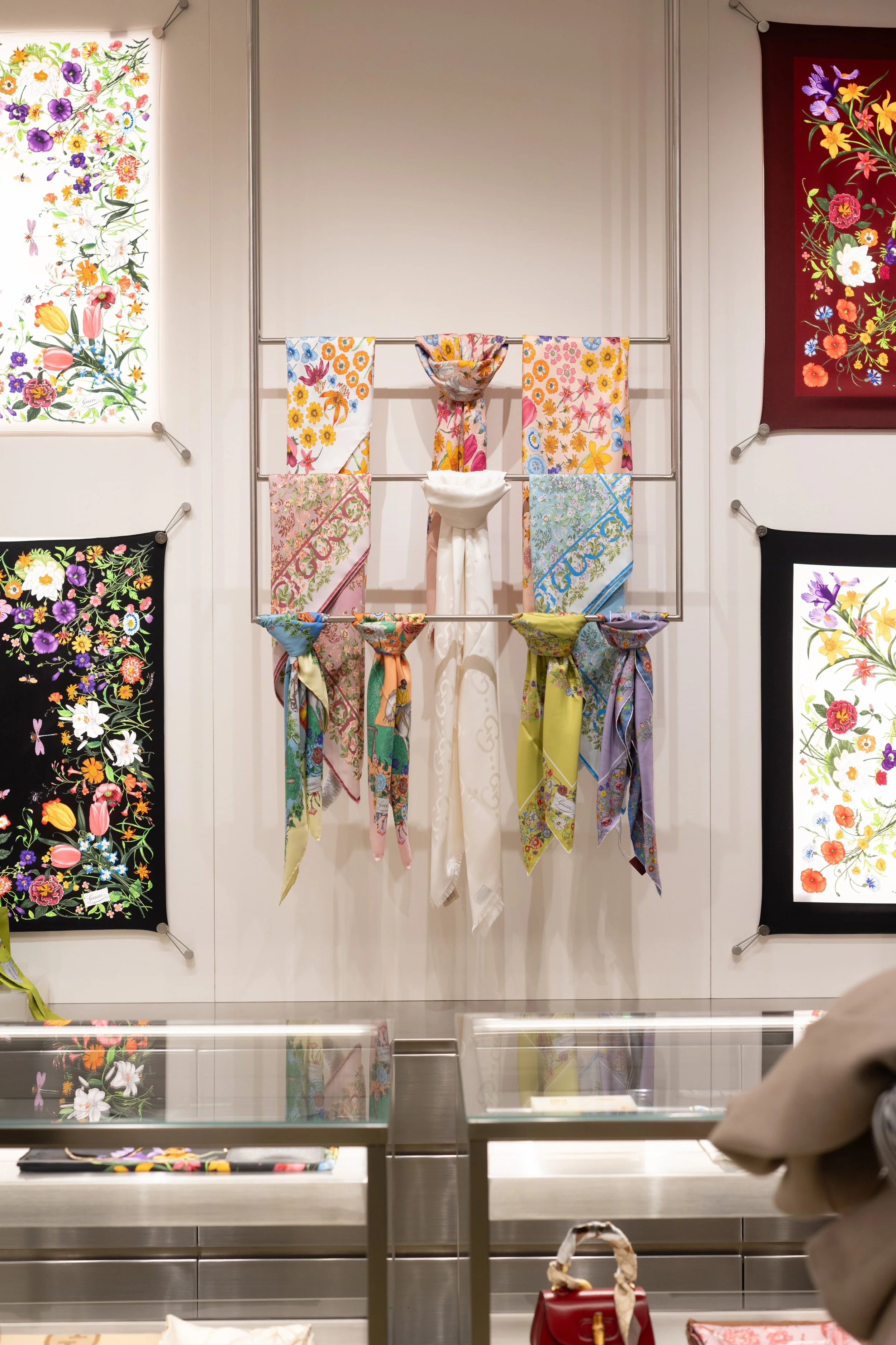 A display of colorful floral scarves hanging on metal rods in a store, with floral artwork on the walls behind them.