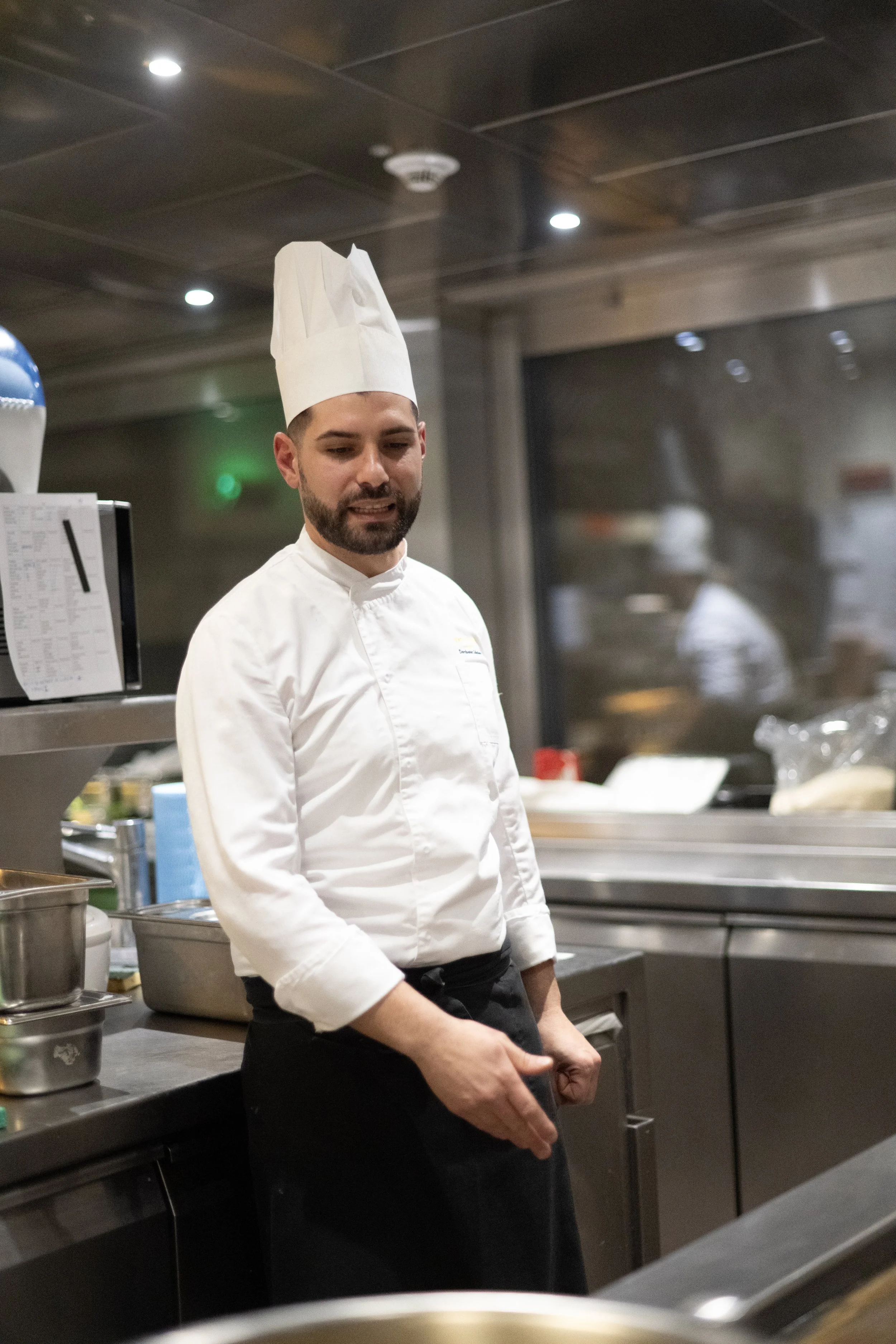 A male chef in a white chef's coat and tall chef hat standing in a professional kitchen.