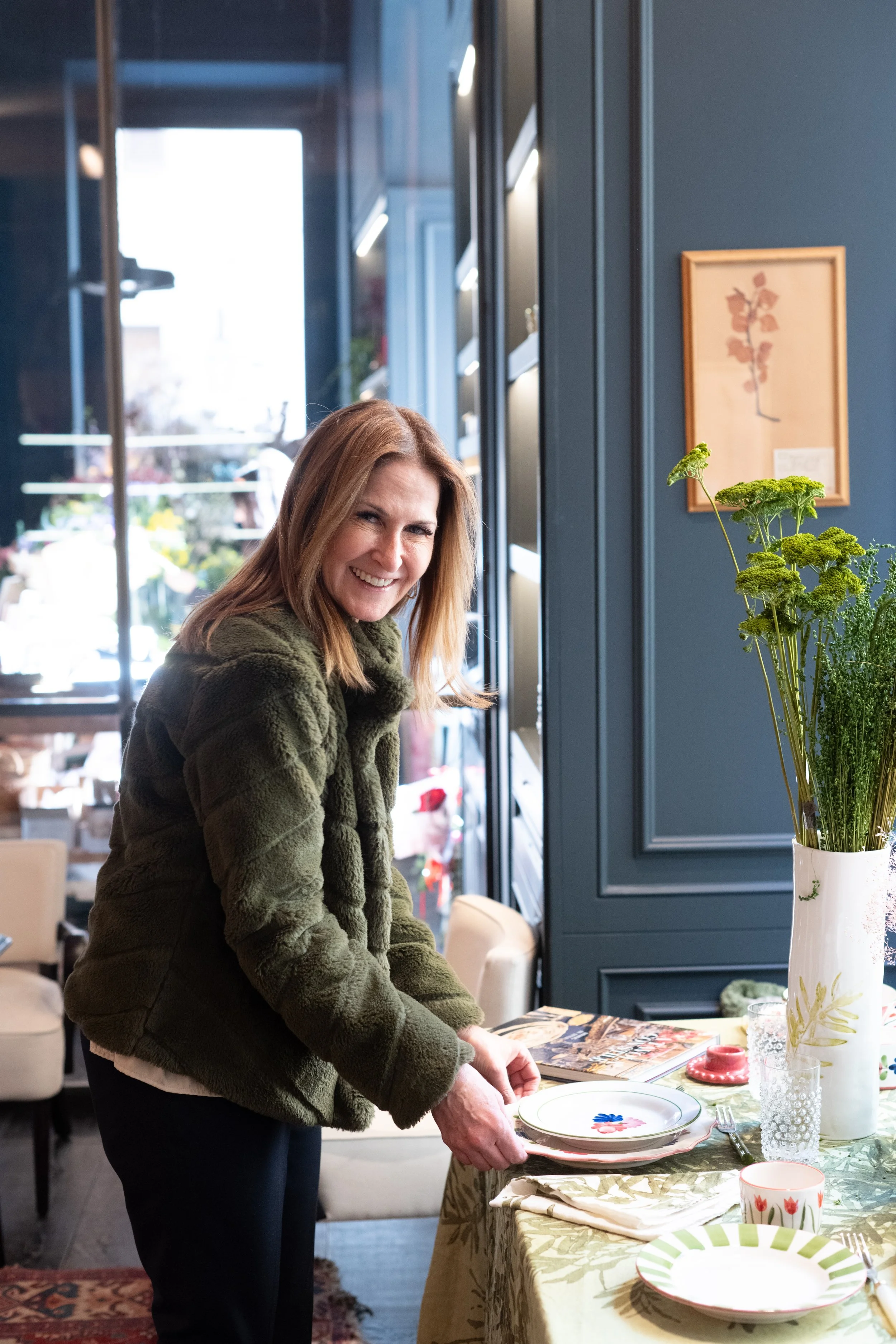 A woman smiling and setting a table in a dining area, with a blue wall, framed artwork, and a vase of green flowers nearby.