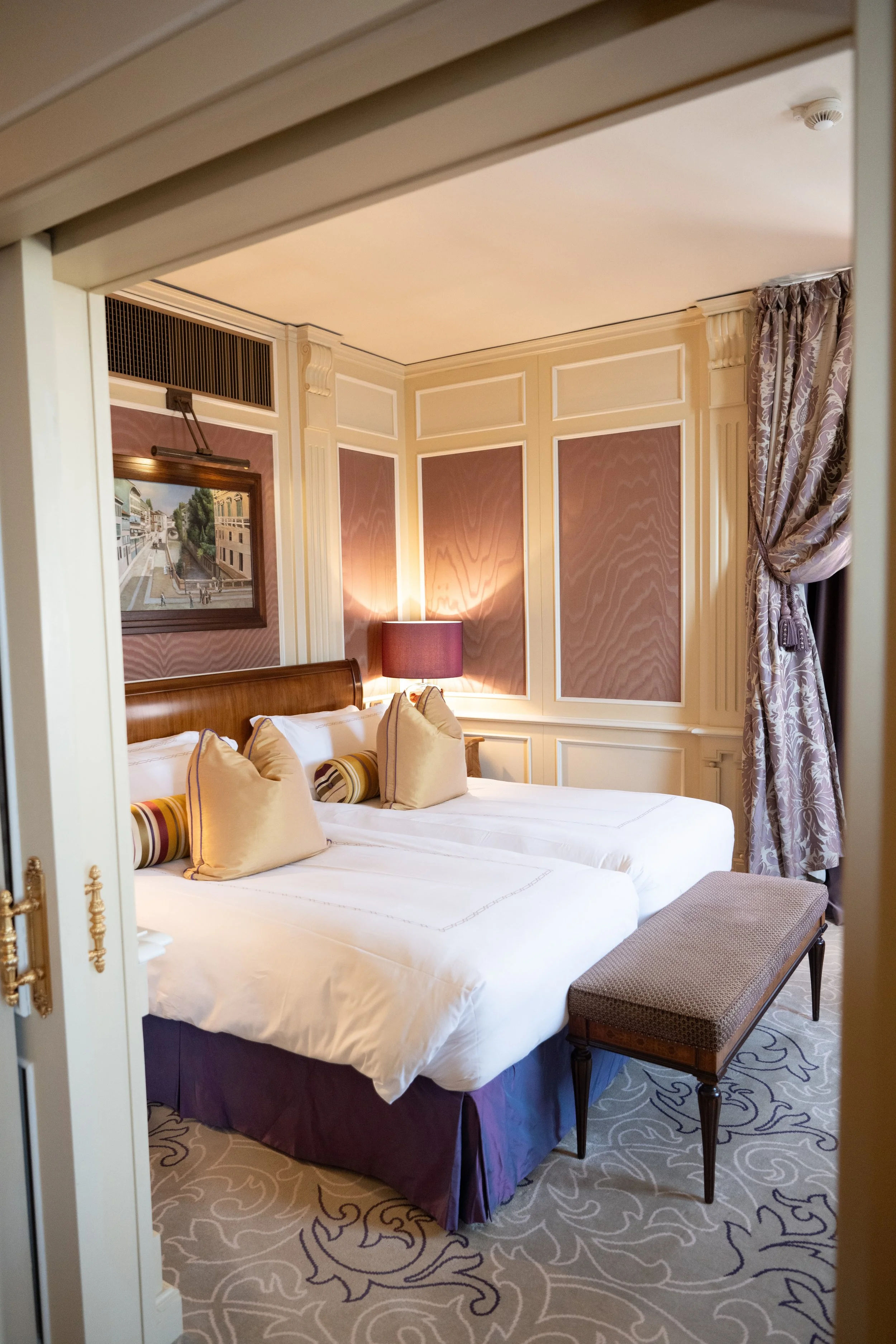 A luxurious hotel room featuring two white beds with pillows, a wooden headboard, a purple bed skirt, and decorative pillows, a patterned carpet, a bench at the foot of the bed, walls with decorative paneling, a framed painting, a table lamp with a p