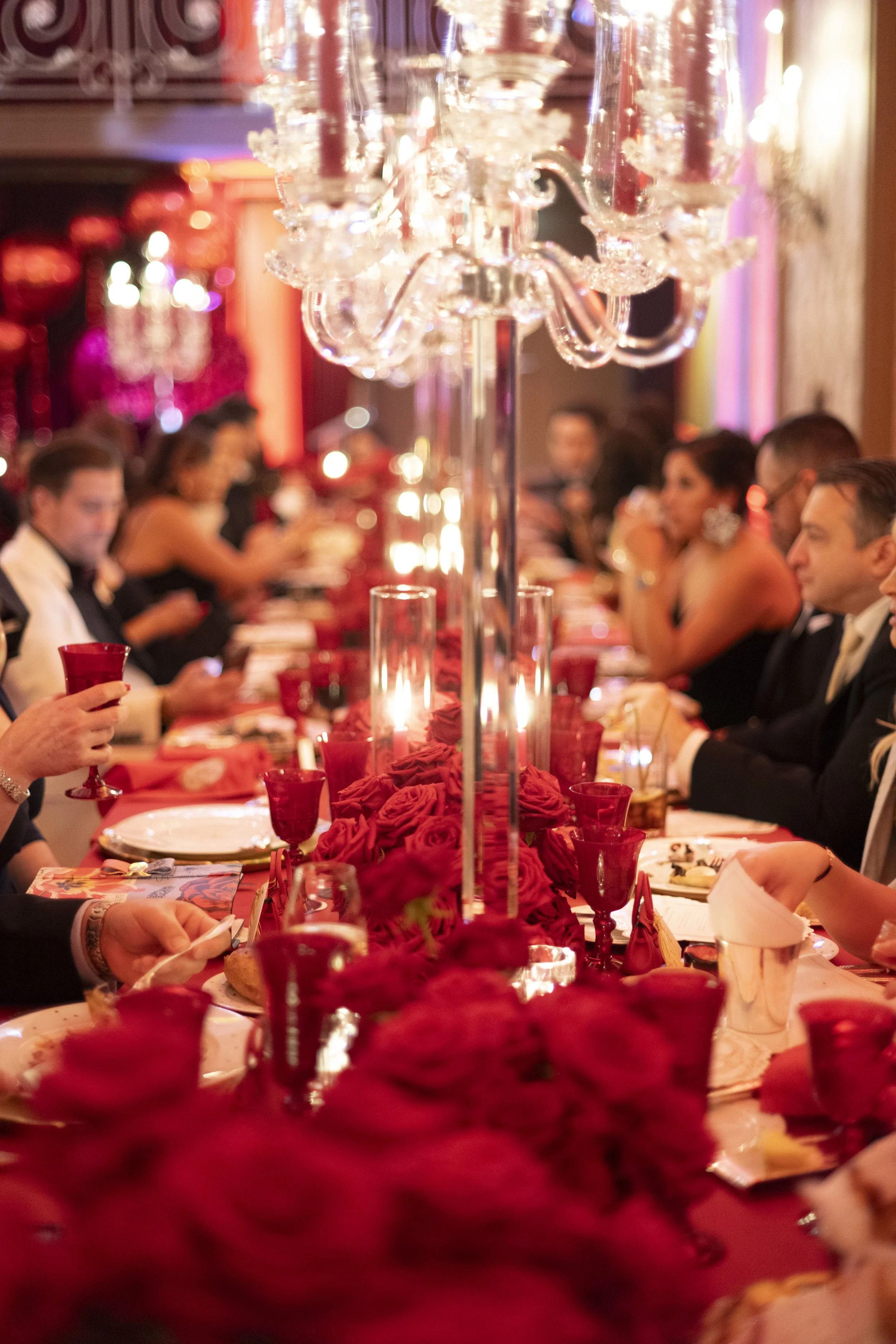 A long banquet table decorated with red roses, red candles, and crystal candle holders, set for a formal dinner. People in formal wear are seated along both sides of the table, engaged in conversation. The background has warm lighting and elegant dec