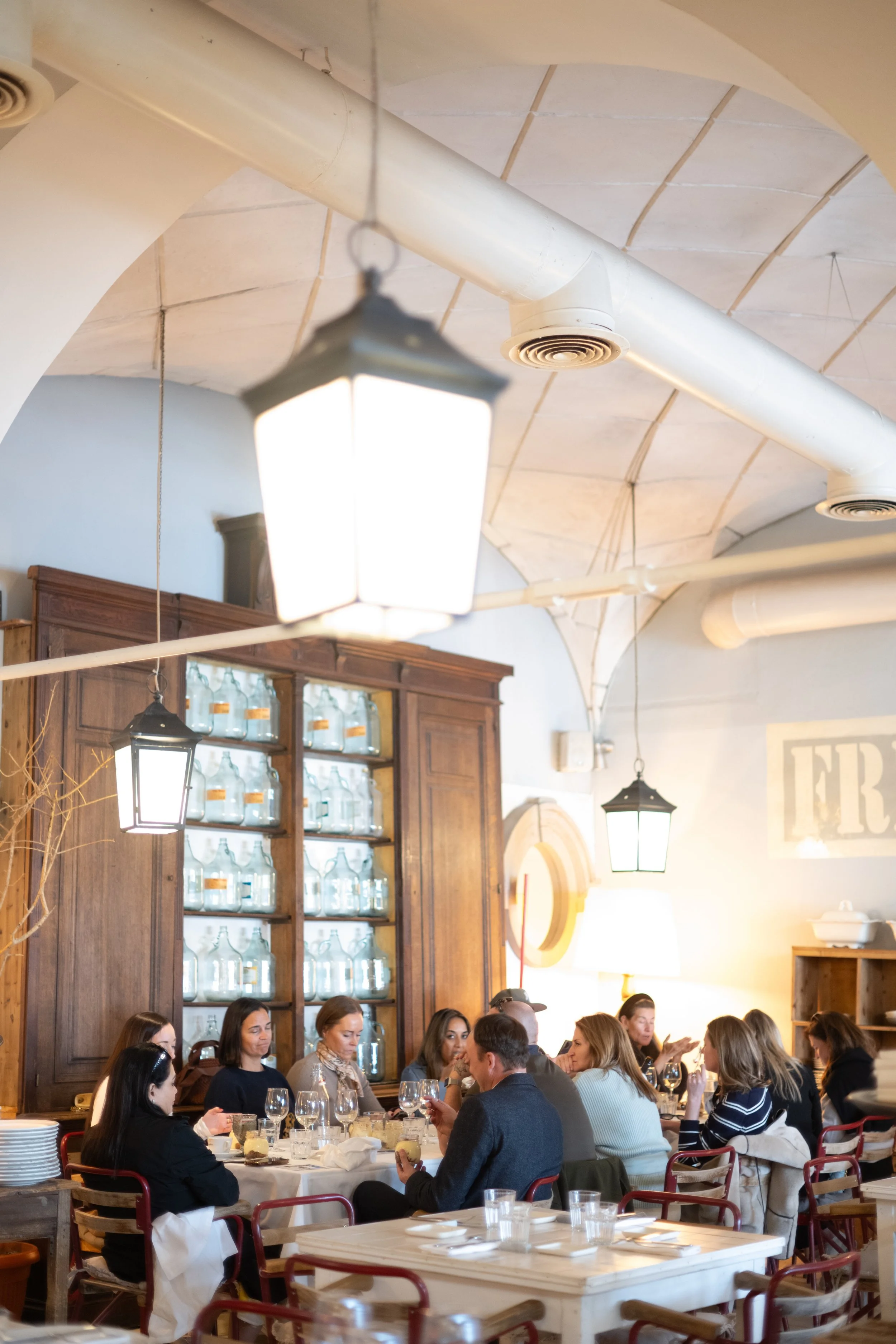 People dining at a restaurant with wooden shelves filled with glass bottles, hanging lantern-style lights, and a vaulted ceiling with exposed ventilation pipes.