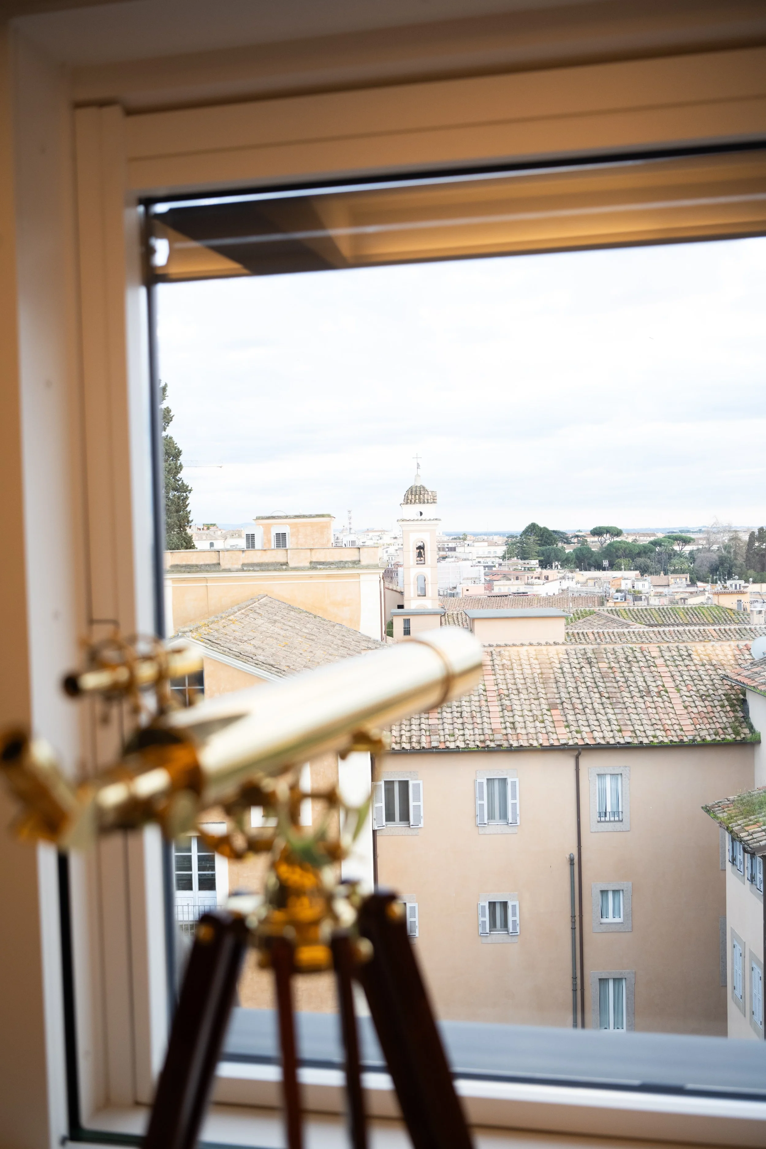 View through a window showing a cityscape with rooftops and a church tower with a bell and a cross, as well as trees and buildings in the background.