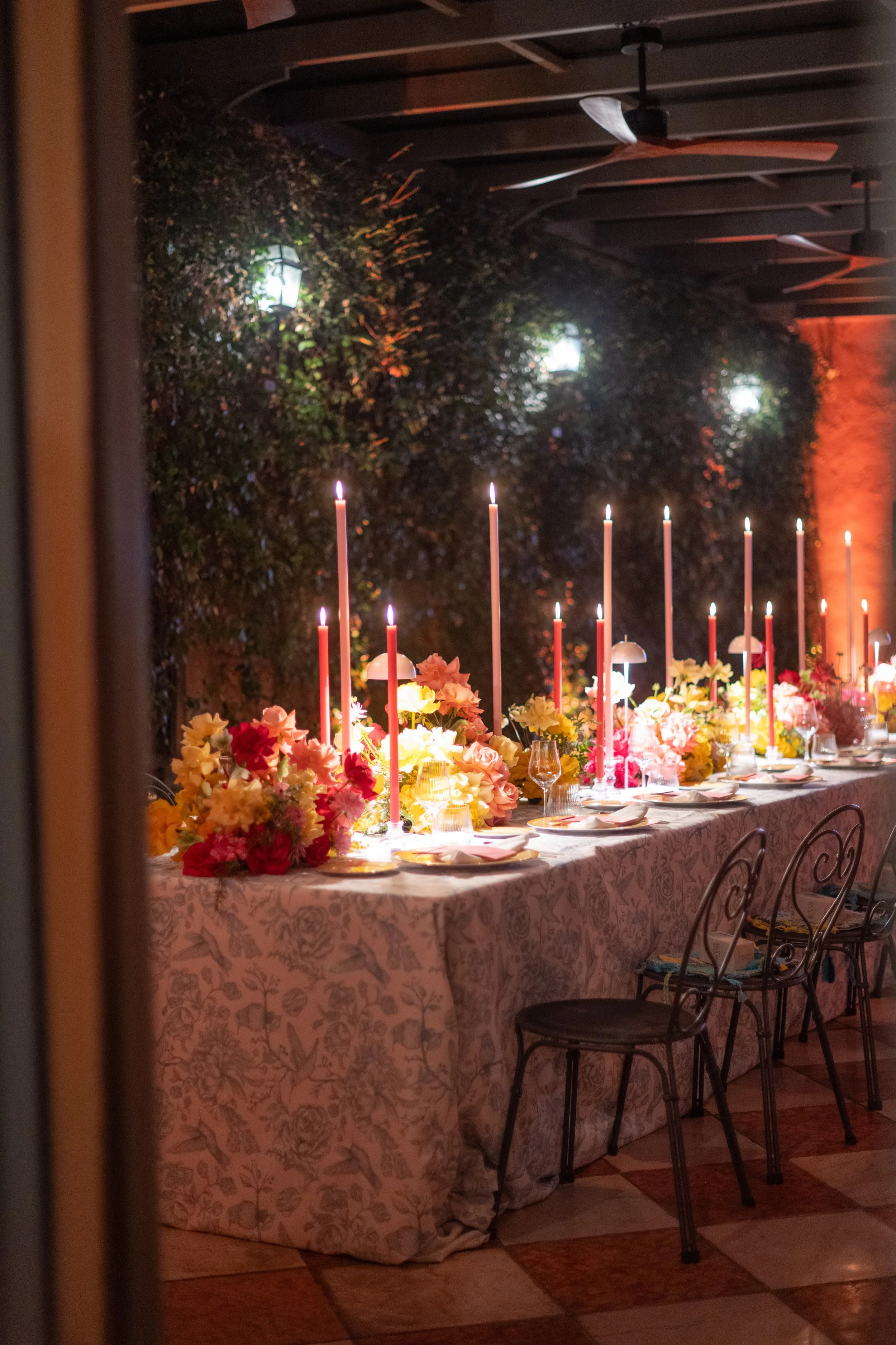 A long dining table is set with floral arrangements and tall pink candles. The room has warm lighting, and a leafy backdrop with hanging lanterns is visible behind the table. A ceiling fan is also in view.