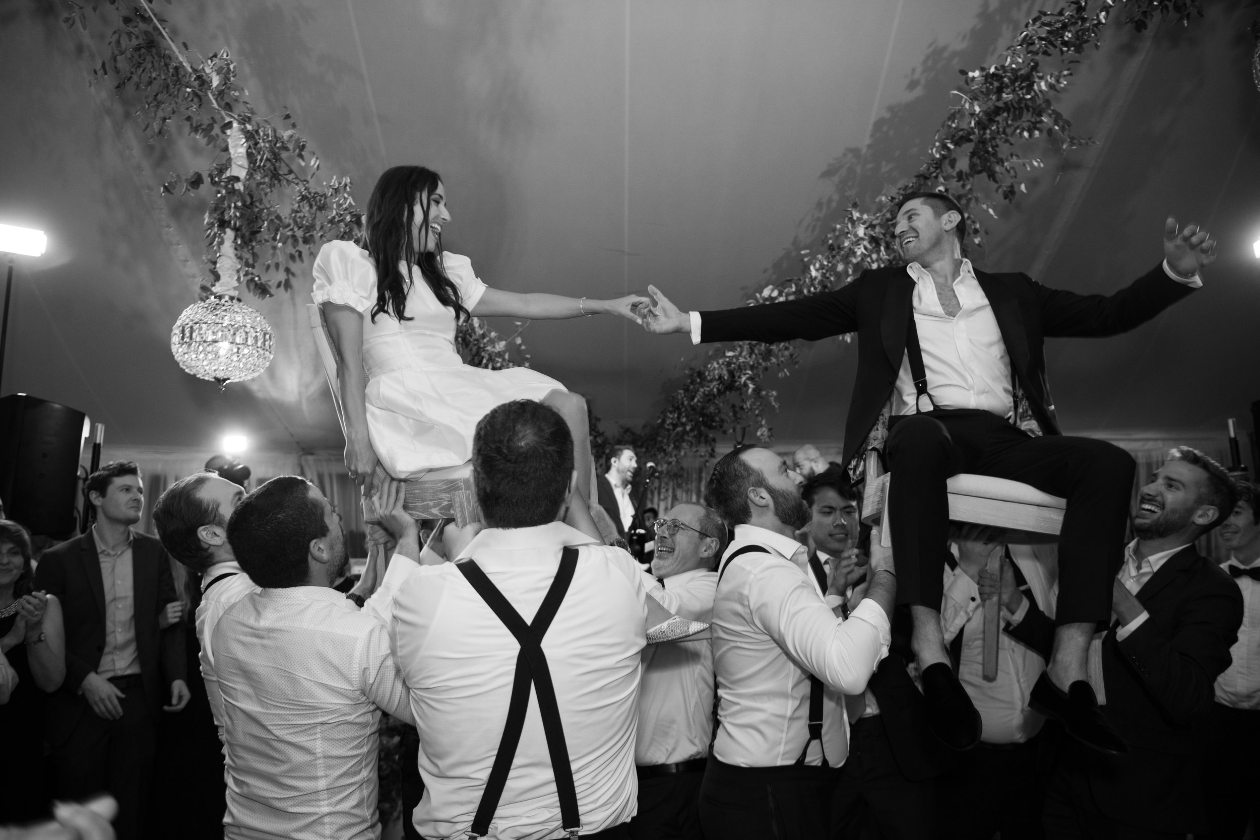 A black-and-white photo of a lively celebration scene with many people. Two individuals are in the center, on pleated chairs, being lifted by others. The woman on the left is wearing a white dress and has long dark hair, reaching out to the smiling m