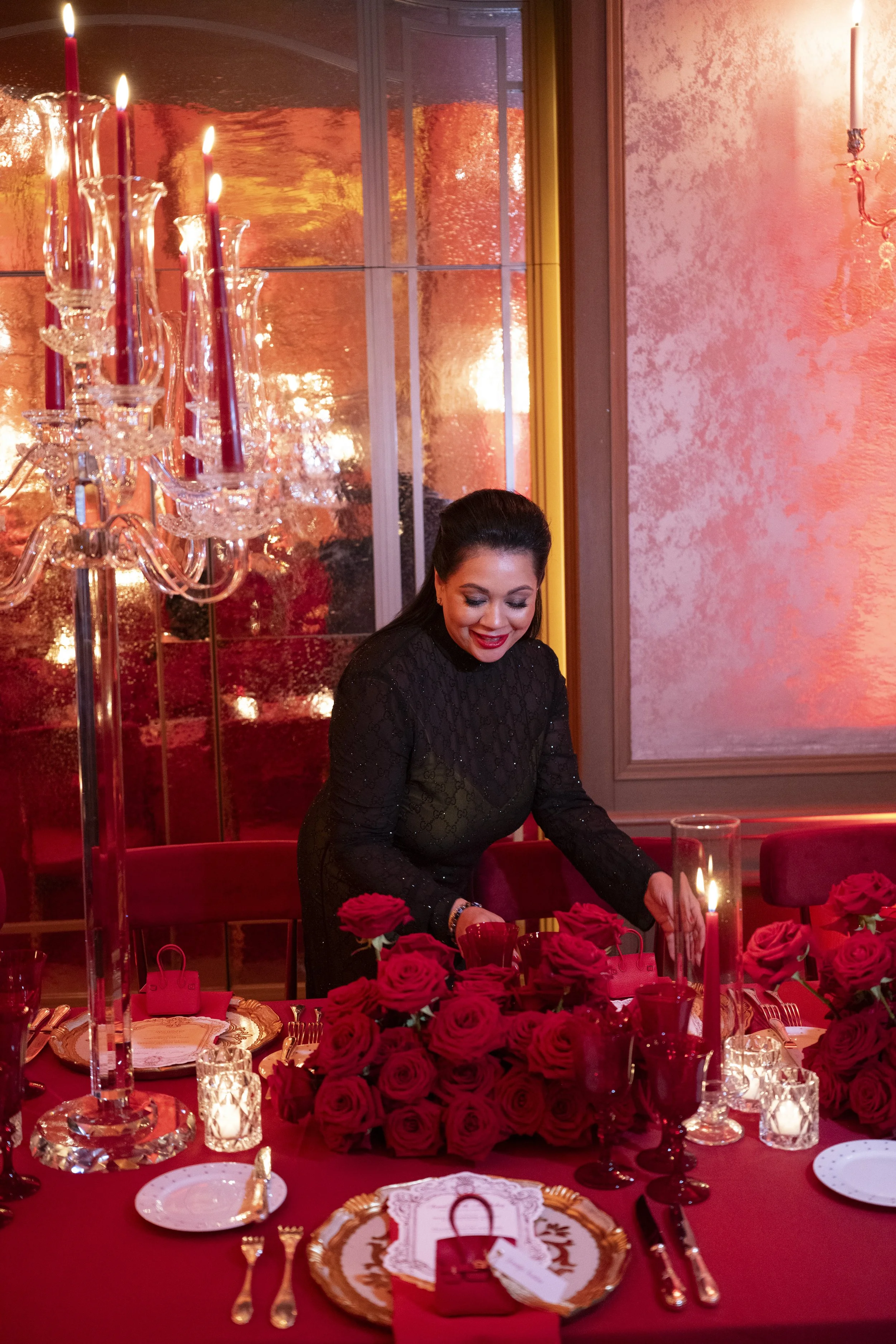 A woman dressed in black is preparing a table set for a dinner party, decorated with red roses, candles, and fine china, in a warmly lit, elegant room with pink textured walls and a large window reflecting warm light.