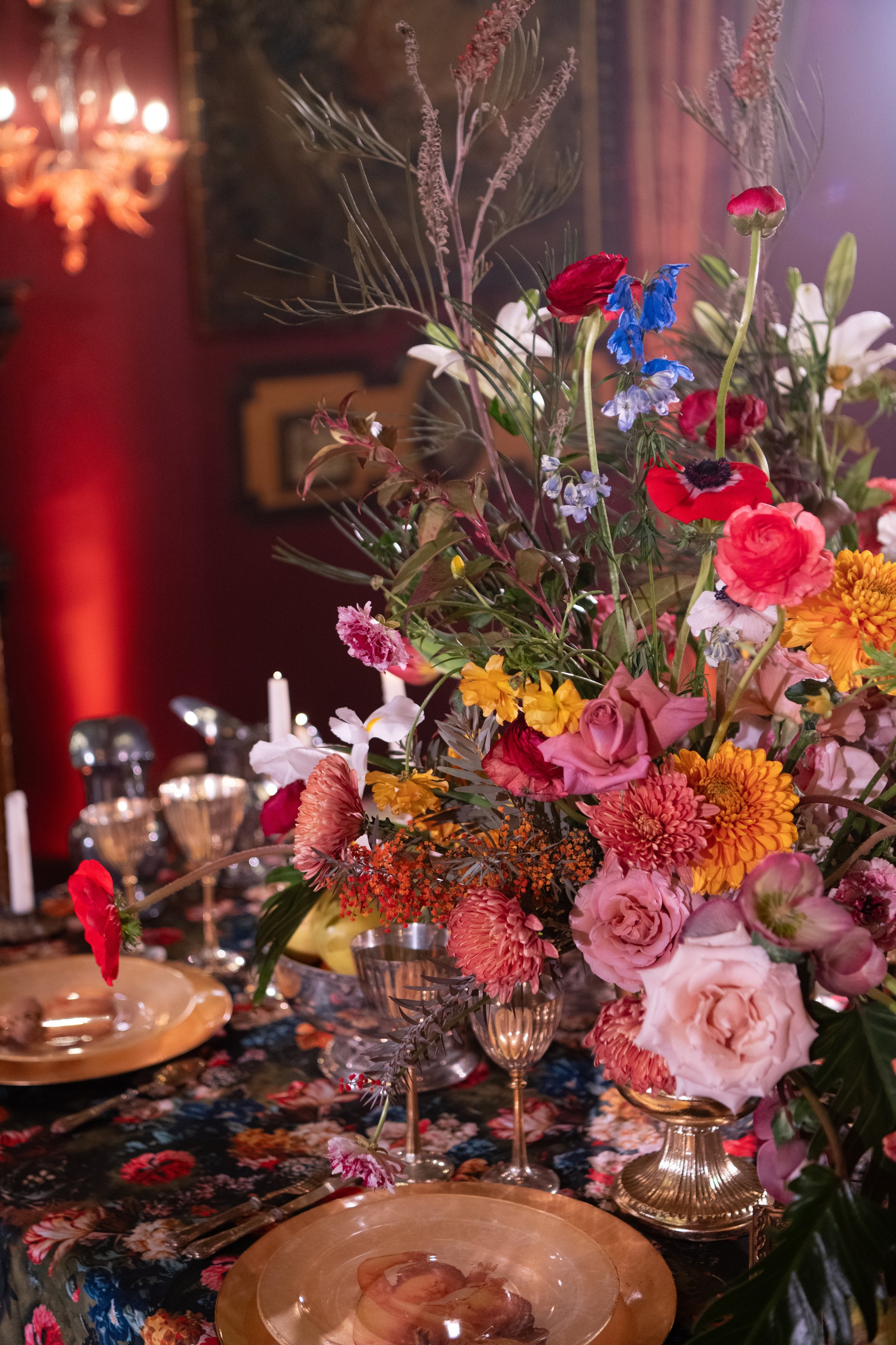 A table set with a large floral arrangement featuring red, pink, white, and blue flowers. The table is covered with a dark floral tablecloth and has gold-rimmed glassware and a gold charger plate with a bread roll.