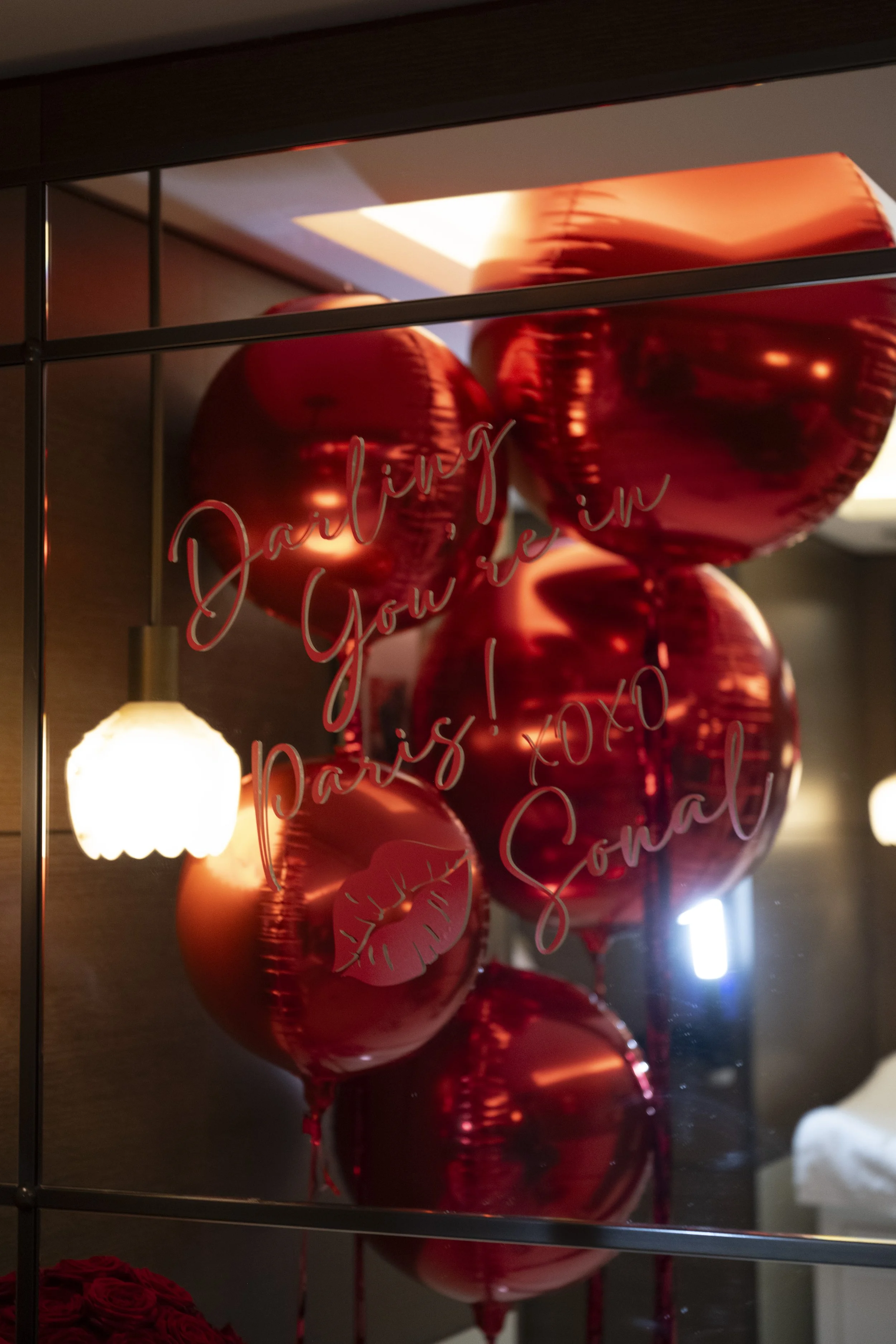 Group of shiny red heart-shaped balloons with the message 'Darling, you are in Paris!'. A small light fixture and some red roses are visible in the background.