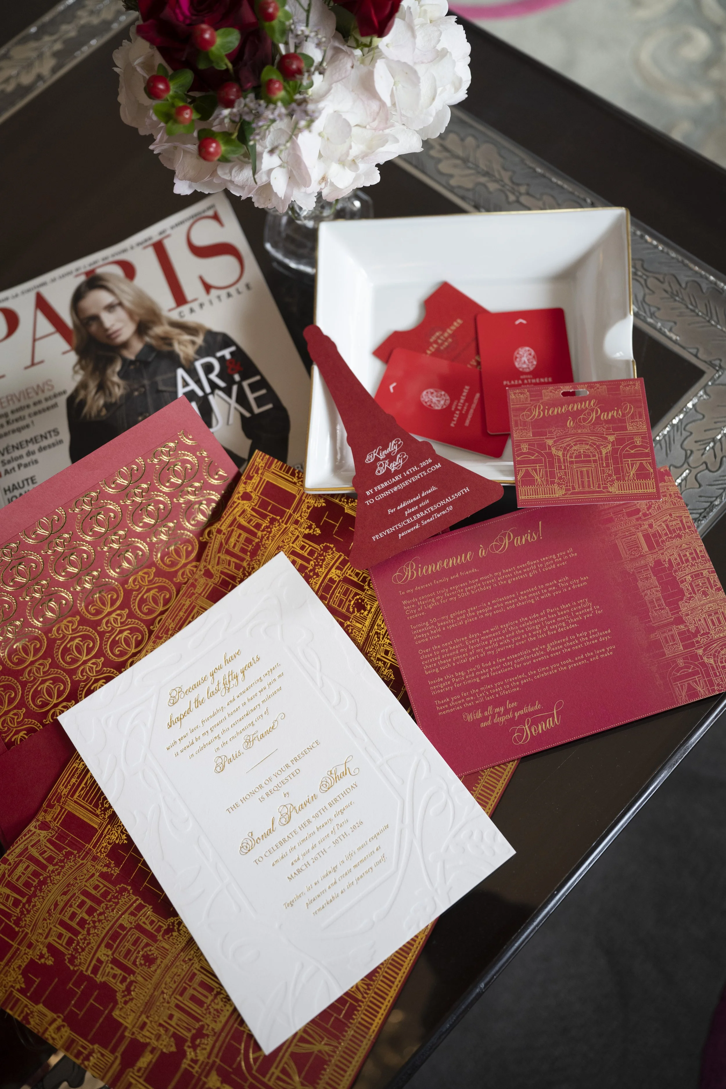 A table with a magazine, a white card with gold decorative embossing, red and gold informational pamphlets, a small white dish, a vase with pink and white flowers, and red cards inside a white dish. The magazine features a woman on the cover and the 