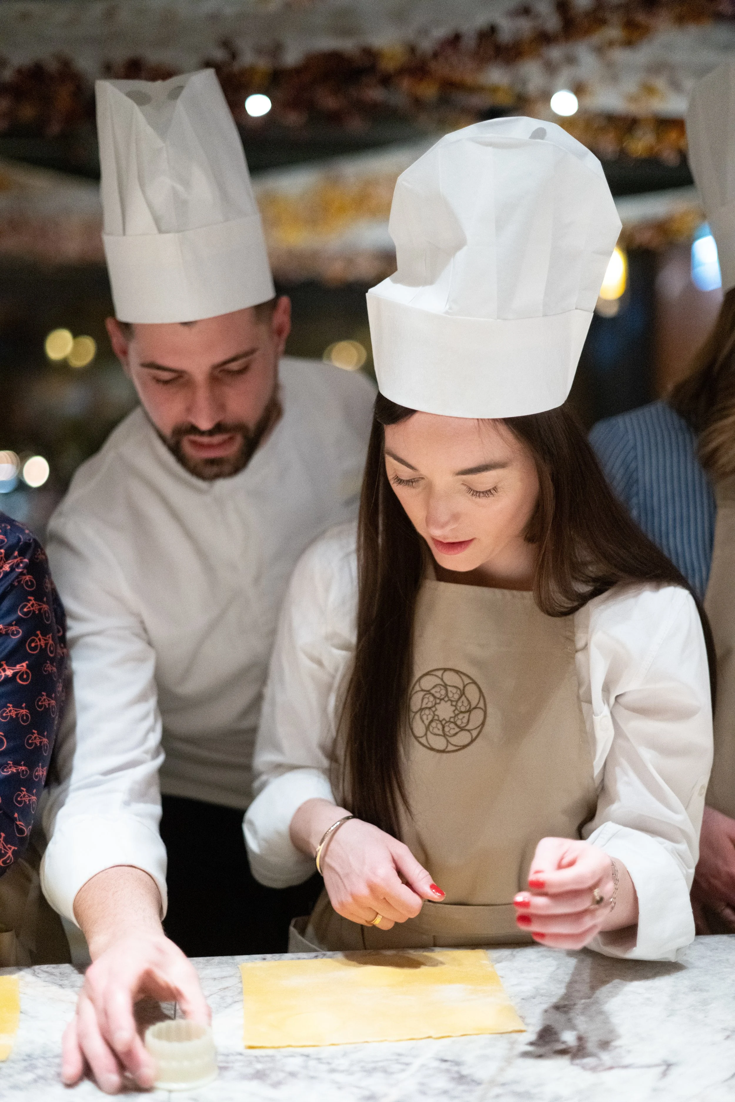 Two people wearing white chef coats and paper chef hats working in a kitchen or restaurant, preparing food on a marble countertop. One person is placing a sheet of pasta or dough, while the other is focused on their task.