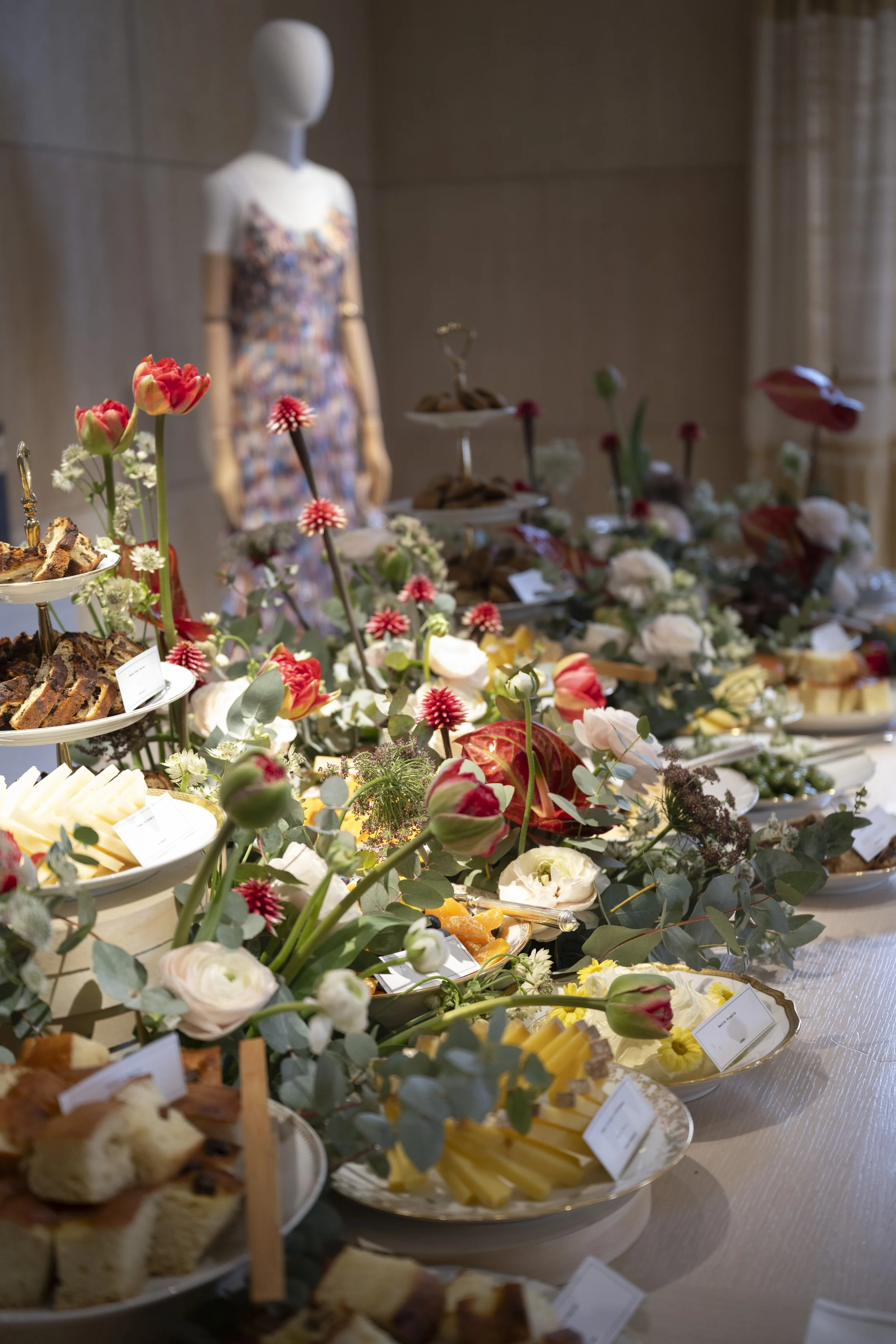 A display table with various desserts and colorful flowers, including sliced cakes, cheese, and decorative flower arrangements, in a well-lit room.