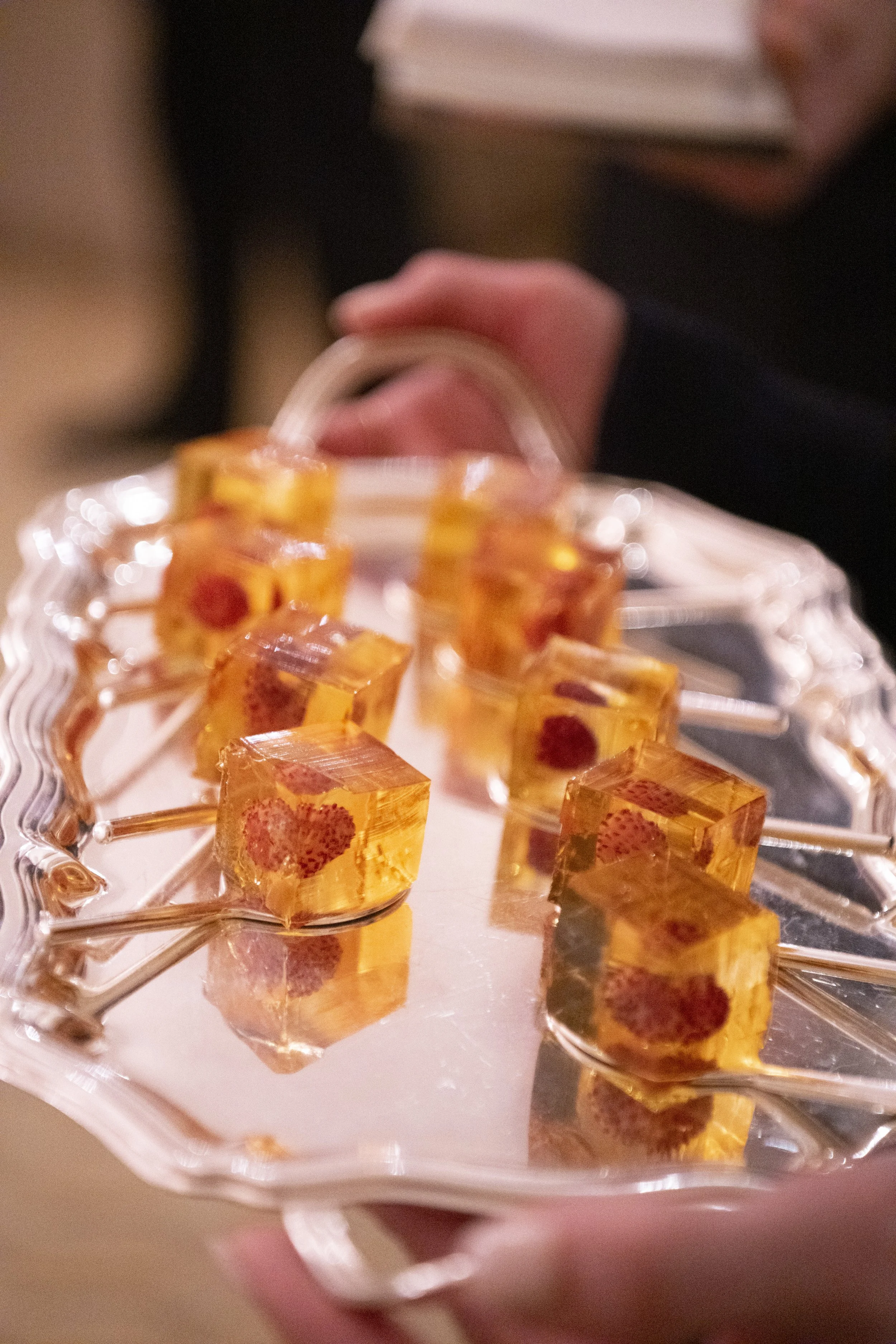 Close-up of a hand holding a tray with square, orange-colored jellies that have red spots inside, arranged on a clear plastic tray.
