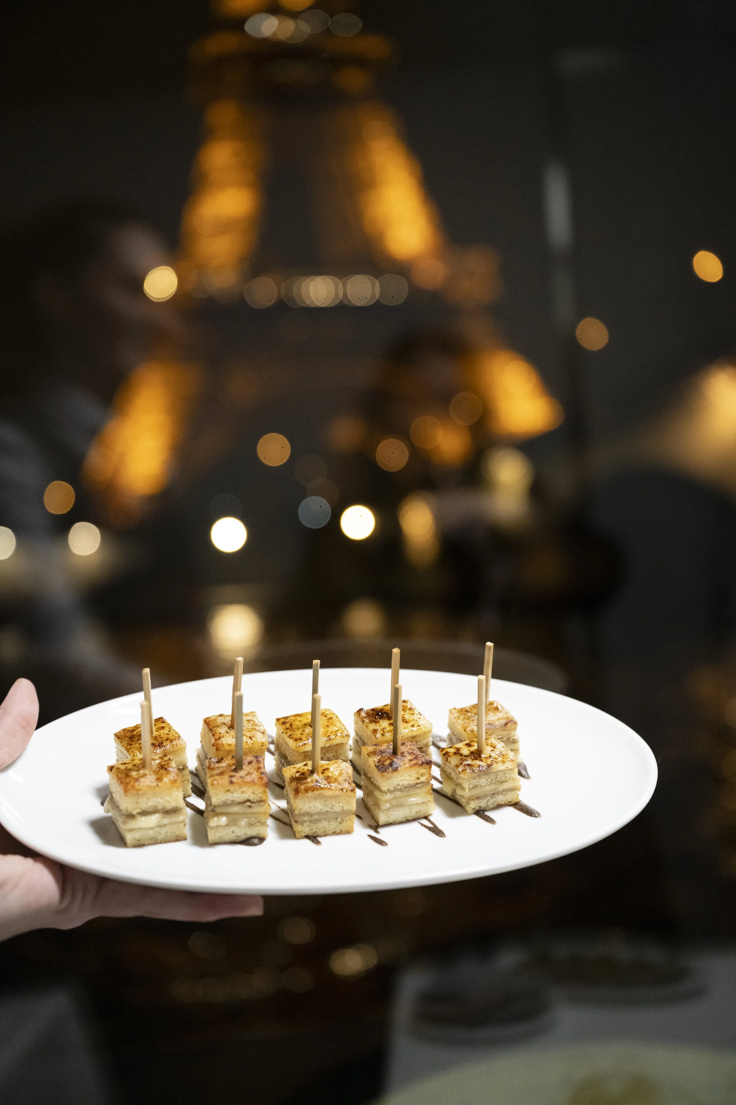 A white plate of small layered food squares with skewers on top, being held by a person, with a blurred cityscape featuring illuminated tall buildings at night in the background.