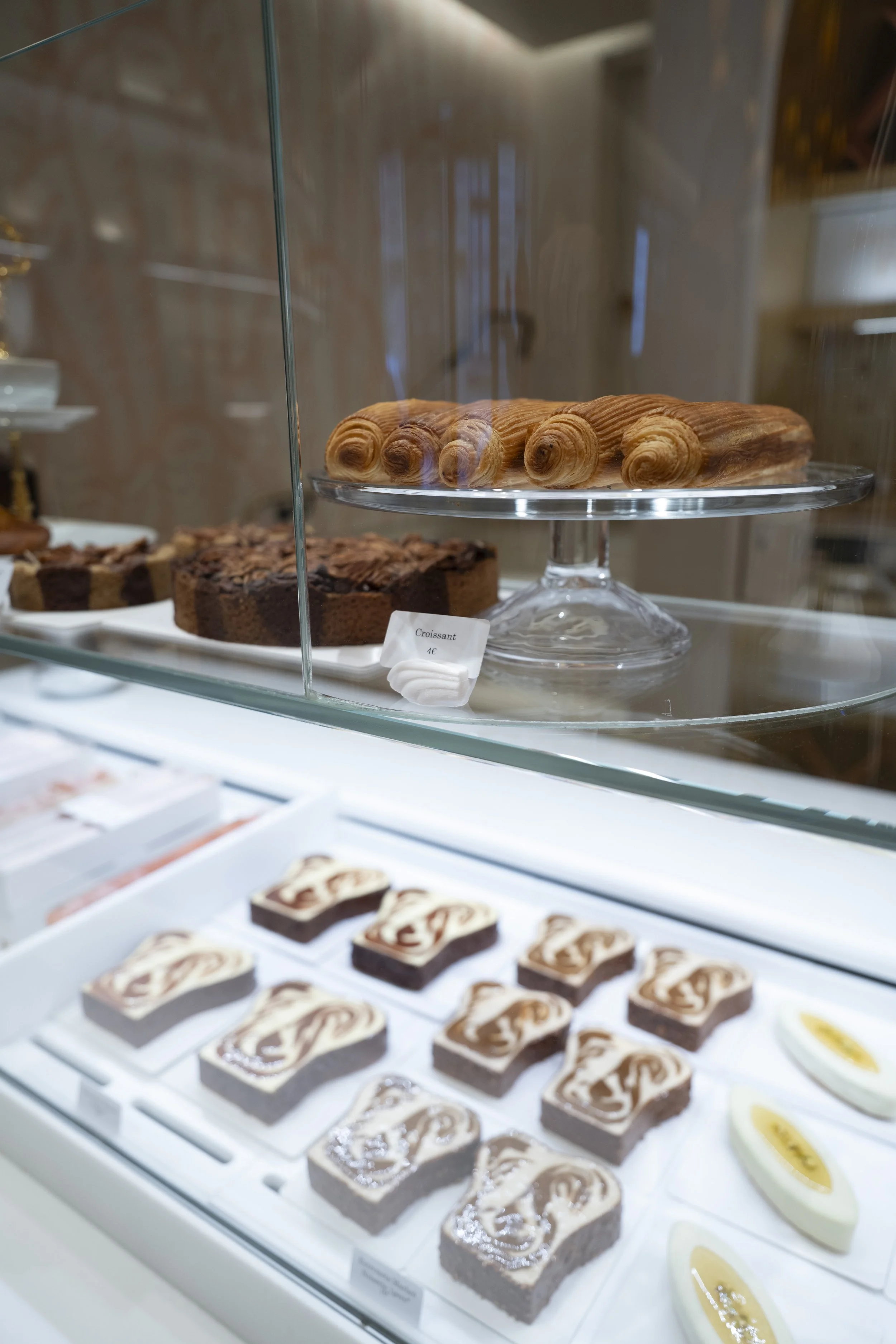 Display case with assorted pastries, including croissants on a glass stand, chocolate cake slices, and marble patterned brownies, in a bakery setting.
