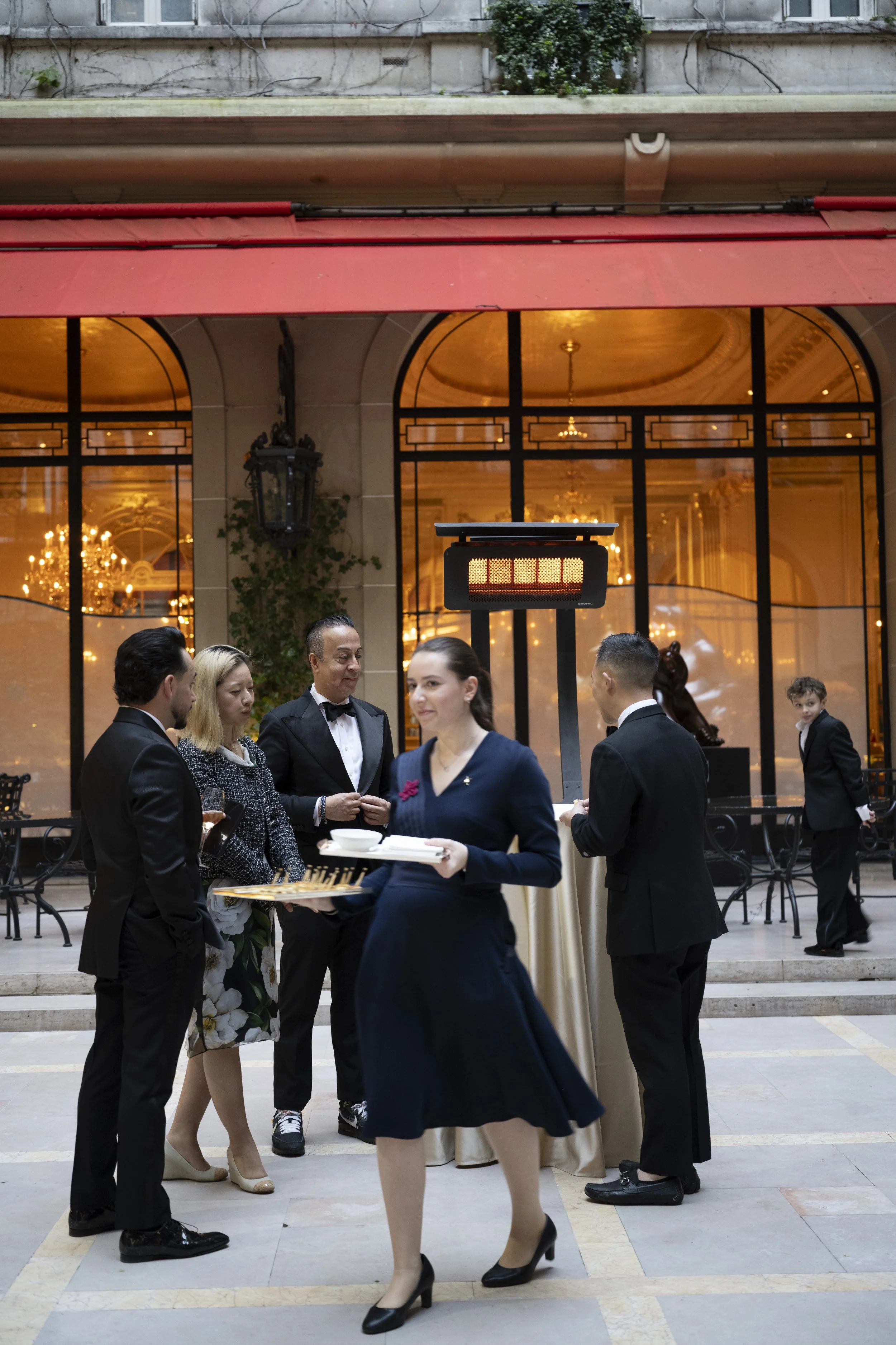 Group of formally dressed people at a social event, standing and conversing inside an elegant building with large glass windows, chandeliers, and ornate interior decor.