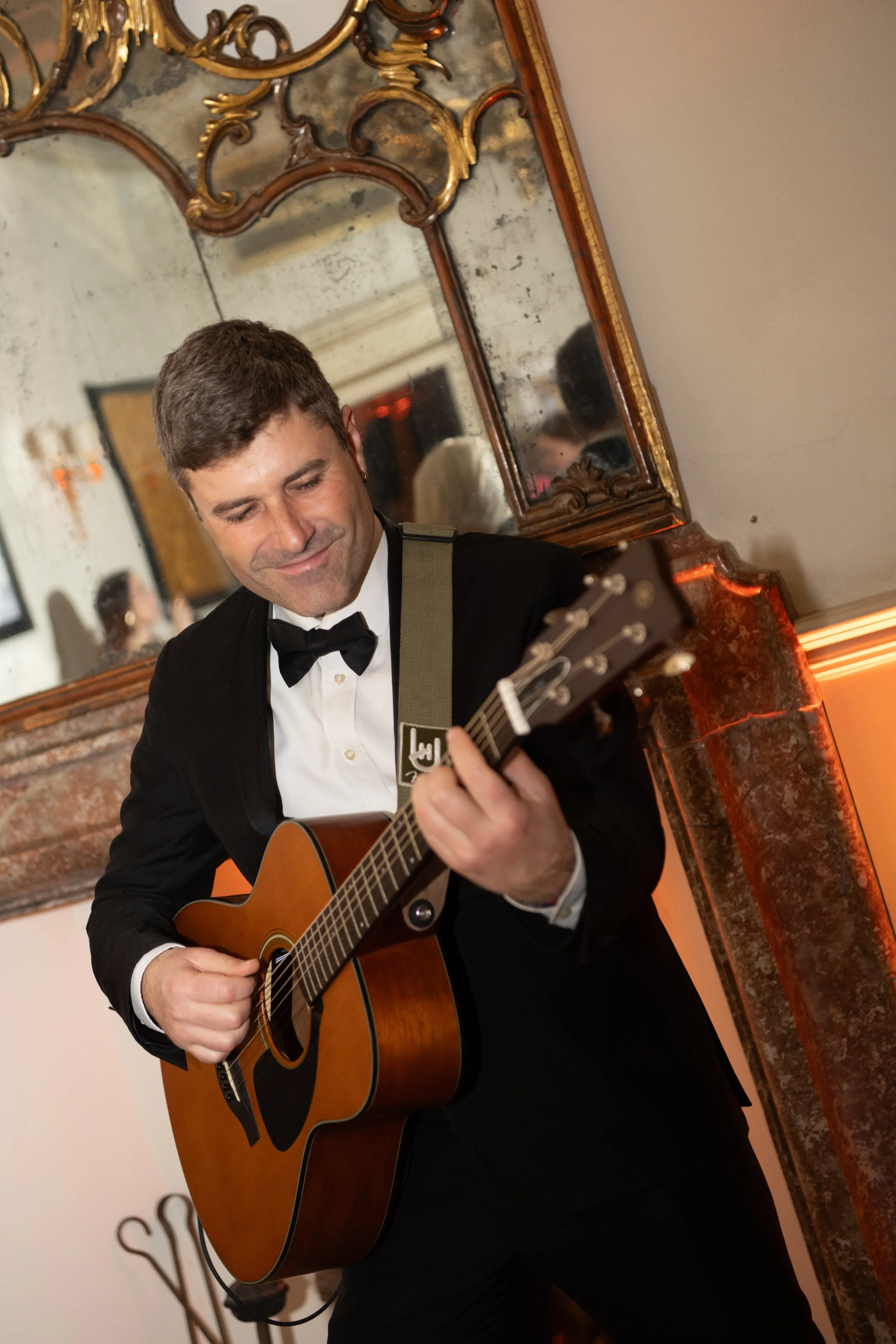A man in a tuxedo playing an acoustic guitar, smiling, standing in front of a vintage mirror and a brick fireplace with a mantle.