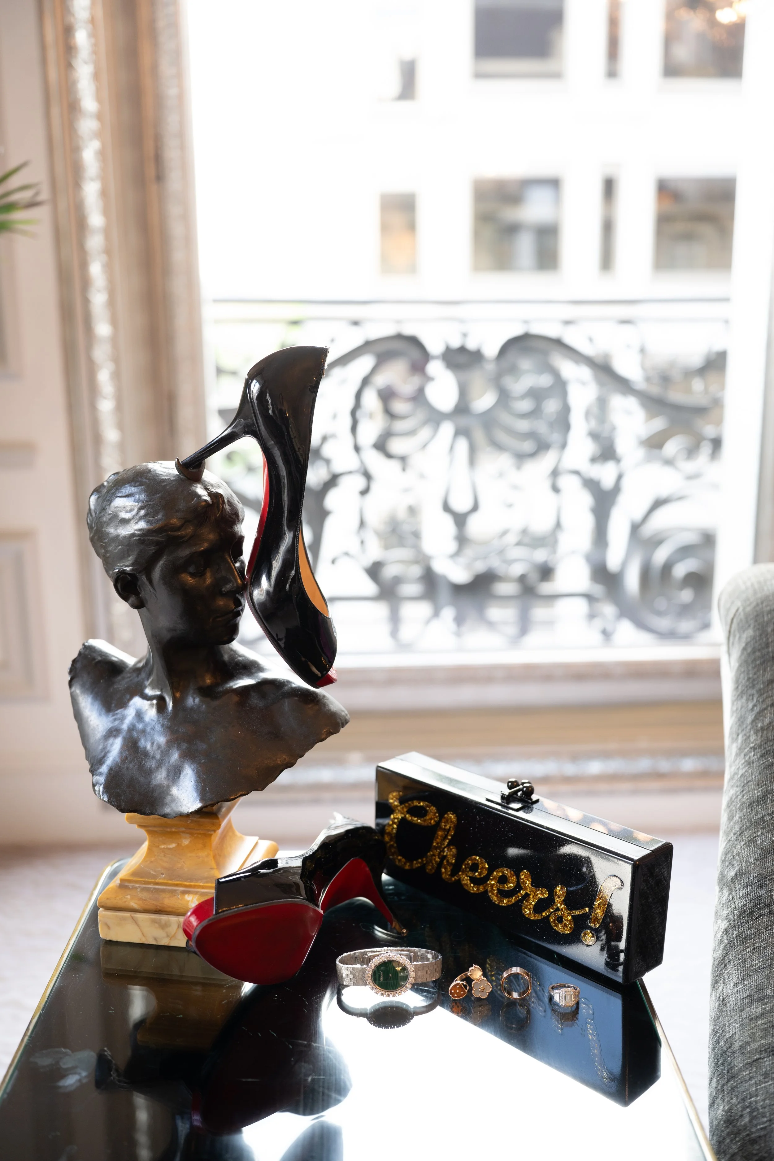 Decorative black and gold bust sculpture of a young man's head with a shoe on top, placed on a marble base. Surrounding objects include a red high-heeled shoe, jewelry, and a black box with gold lettering that reads 'cheers!' with a background of a b