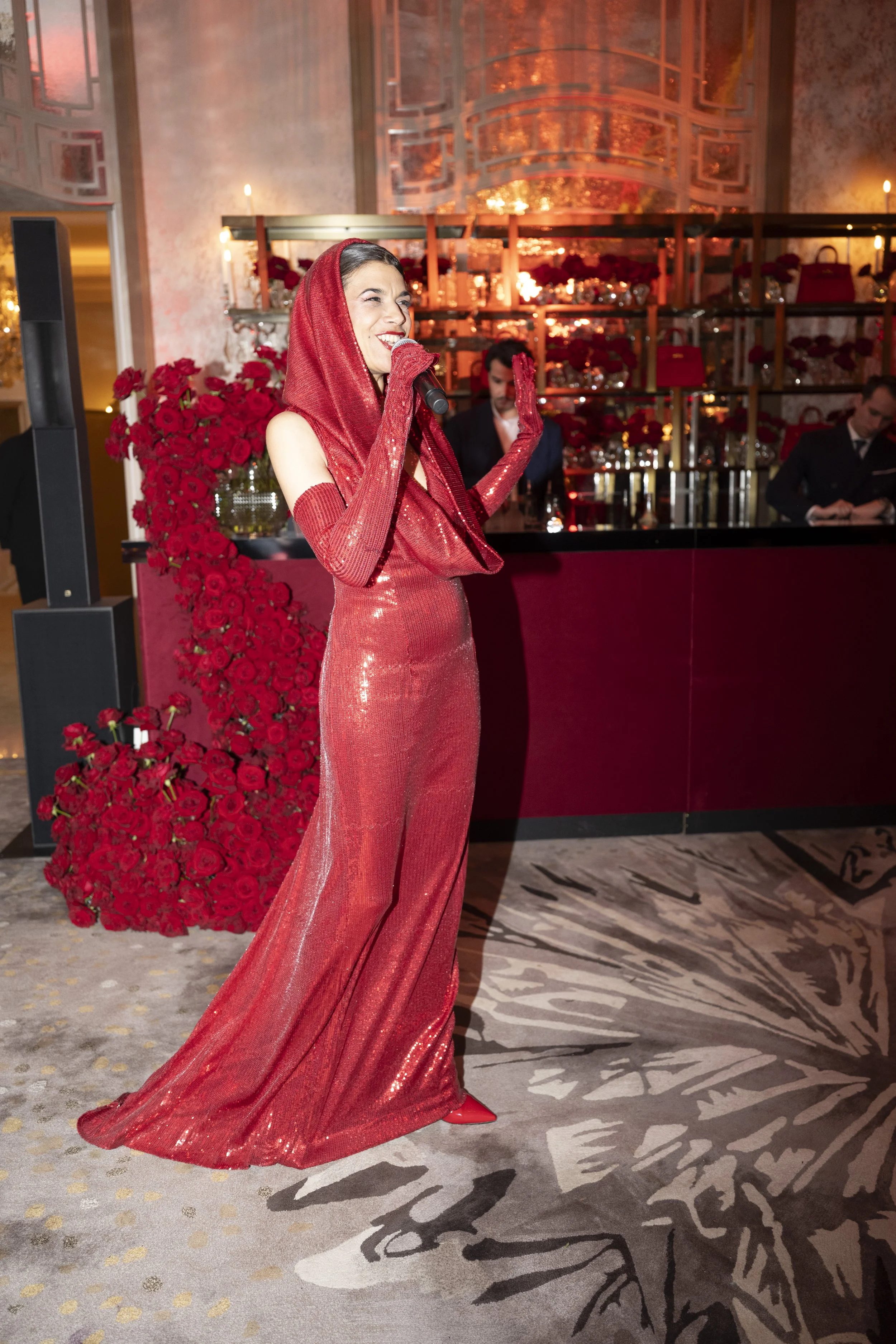 A woman in a red, sequined, floor-length gown with a matching head covering, standing in front of a decorated background with roses and a bar, holding a microphone and smiling.