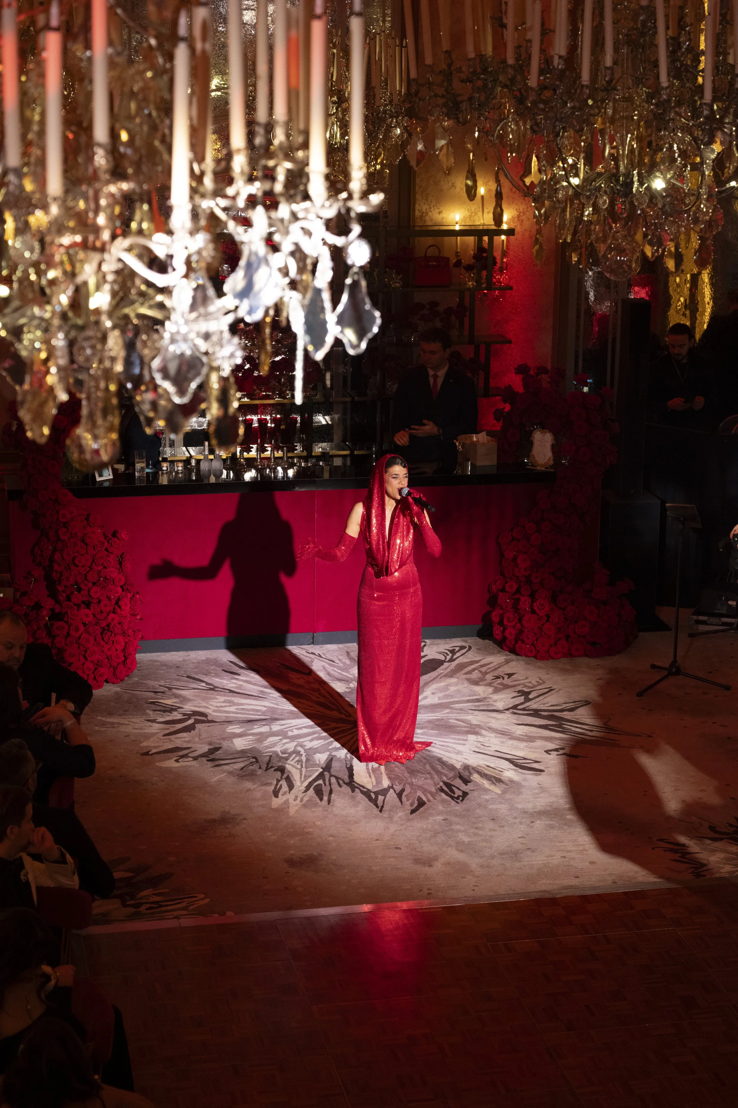 A female singer in a red gown performs on stage, illuminated by spotlight, with ornate chandeliers and a velvet backdrop in the background.