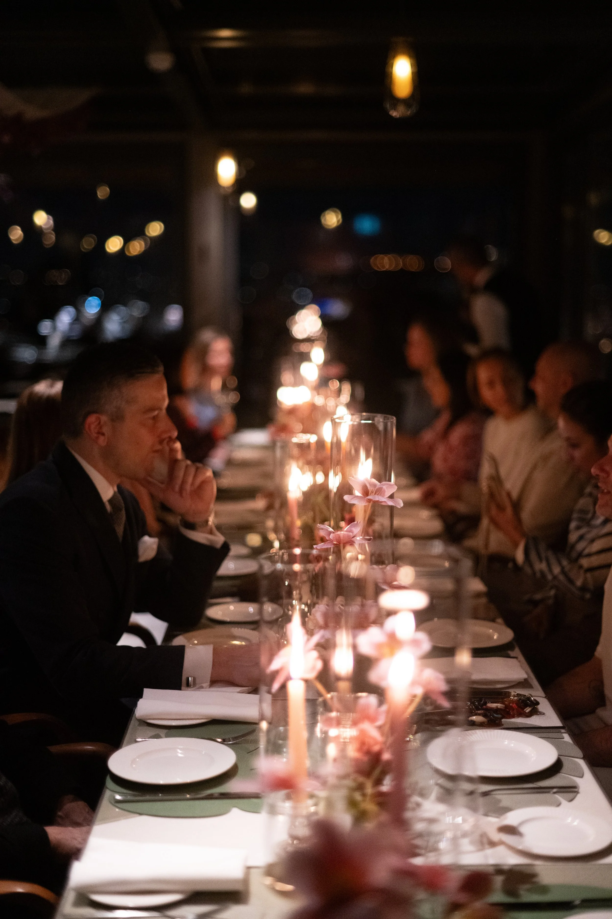 People gathered around a long dinner table at night, with candlelight and hanging lights creating a warm ambiance.