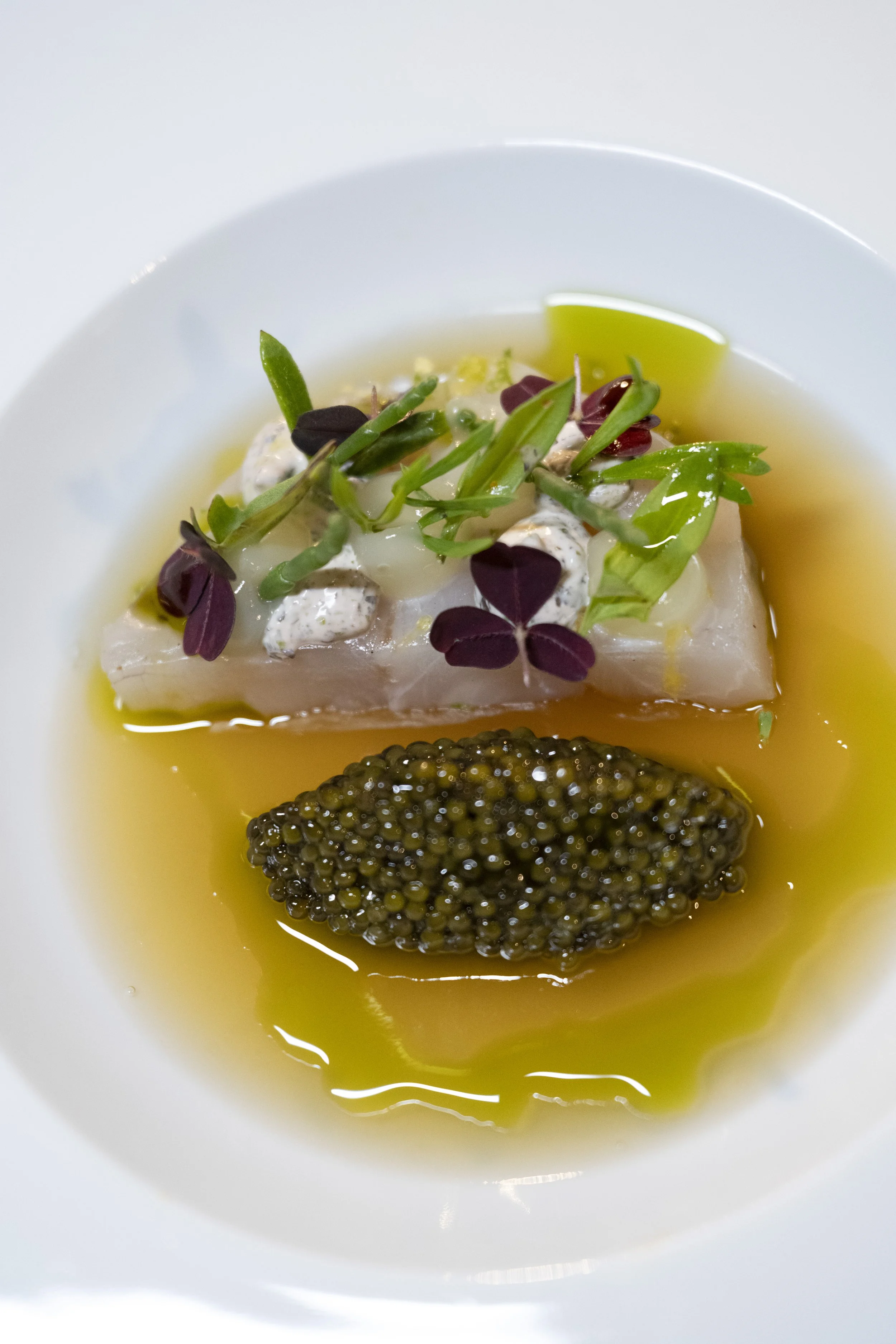 Close-up of a gourmet dish with a piece of fish topped with microgreens and creamy sauce, served in a yellow oil base with a mound of black caviar on the side, in a white bowl.