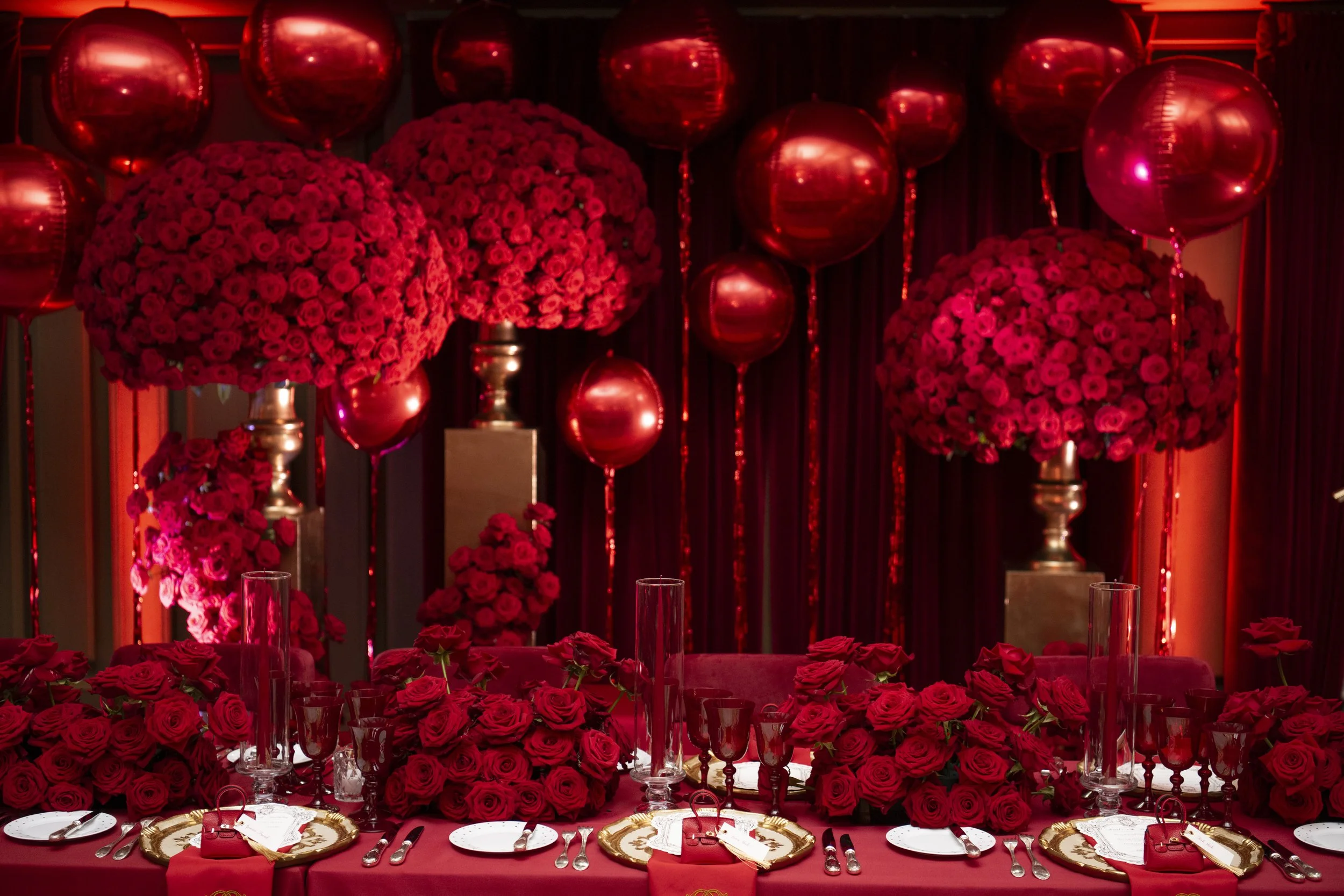 A table decorated with numerous red roses, surrounded by glassware, plates, and cutlery, with red balloons and large rose arrangements in the background.