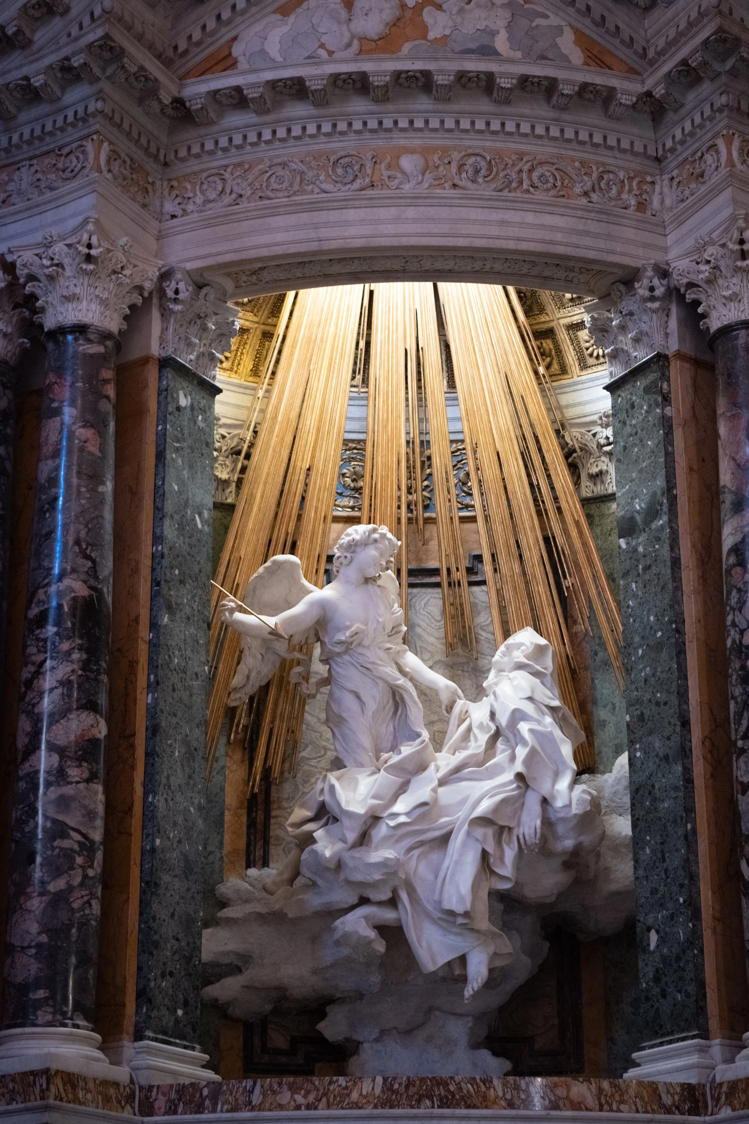 A detailed marble and gold sculpture with intricately carved architectural elements and classical motifs, featuring a central dynamic white marble statue of a mythological figure surrounded by hanging golden rods or beams.