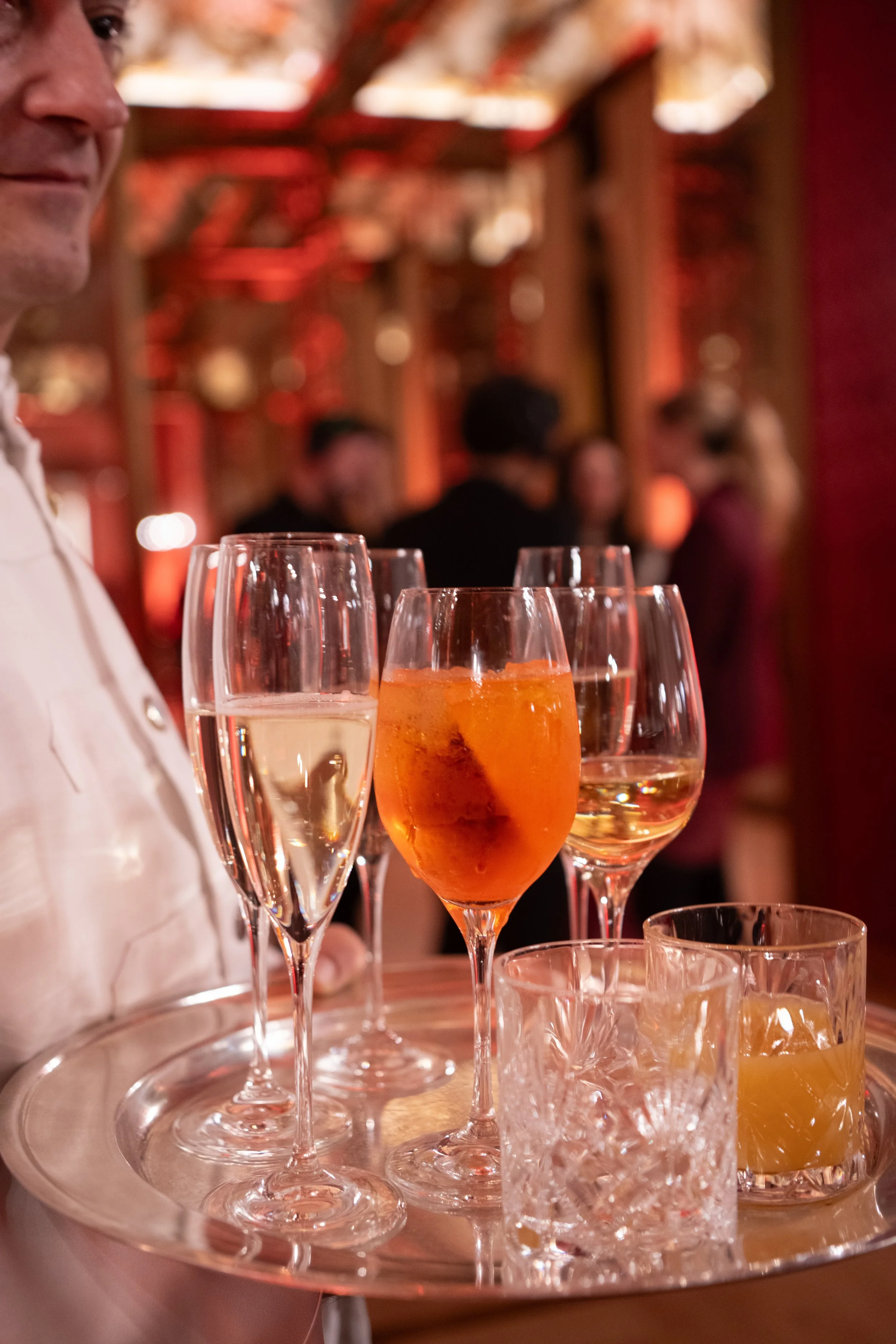 Close-up of colorful champagne glasses on a silver tray at an indoor party, with a blurred background of people and warm lighting.