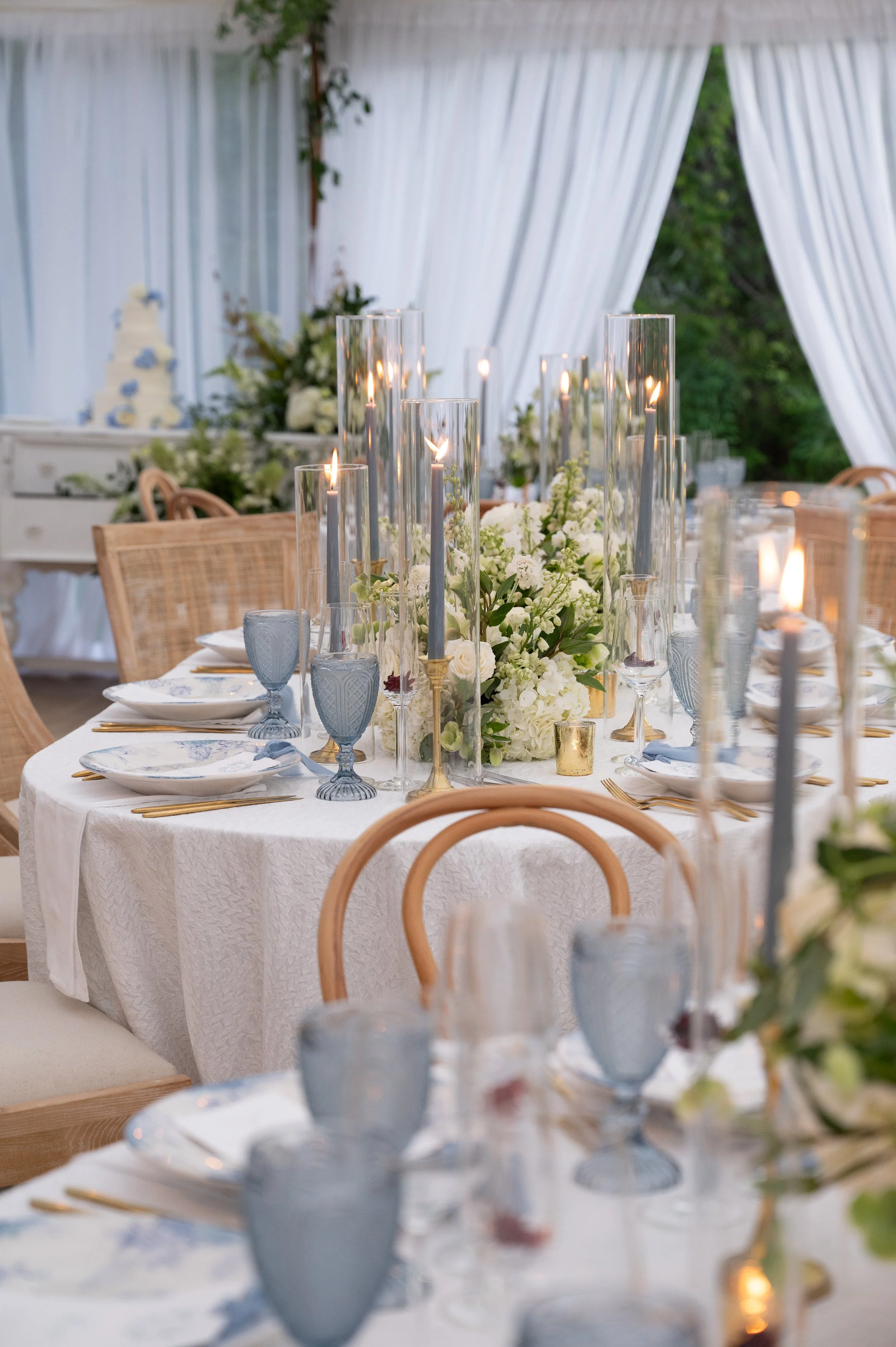 Elegant dining table setup with white tablecloth, tall glass candle holders with lit candles, blue and clear glassware, white and gold dinnerware, and floral centerpieces in white and green, under white drapes.