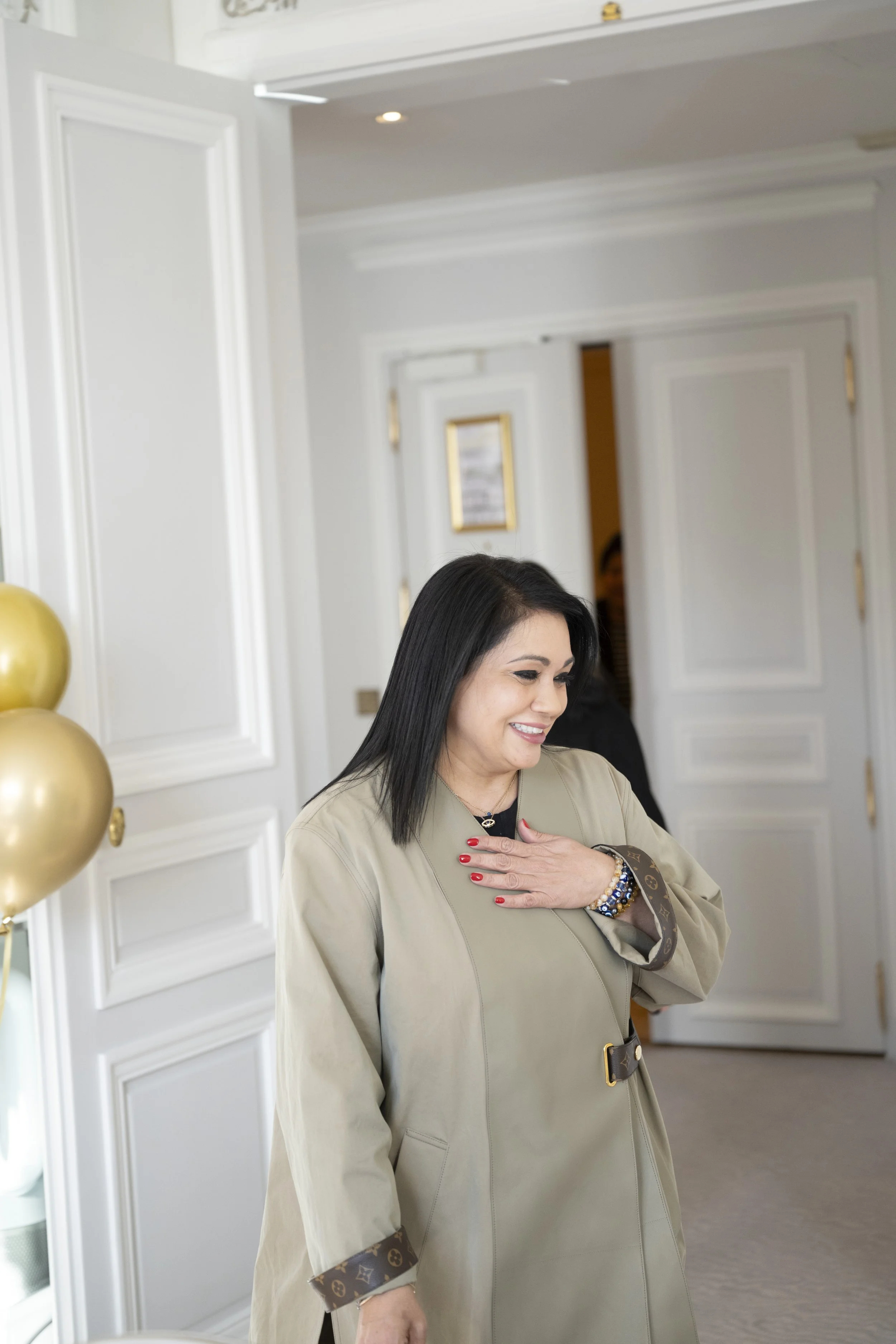 A woman with black hair, wearing a beige coat with designer trim, smiling and placing her hand over her heart indoors near white paneled doors and gold balloons.