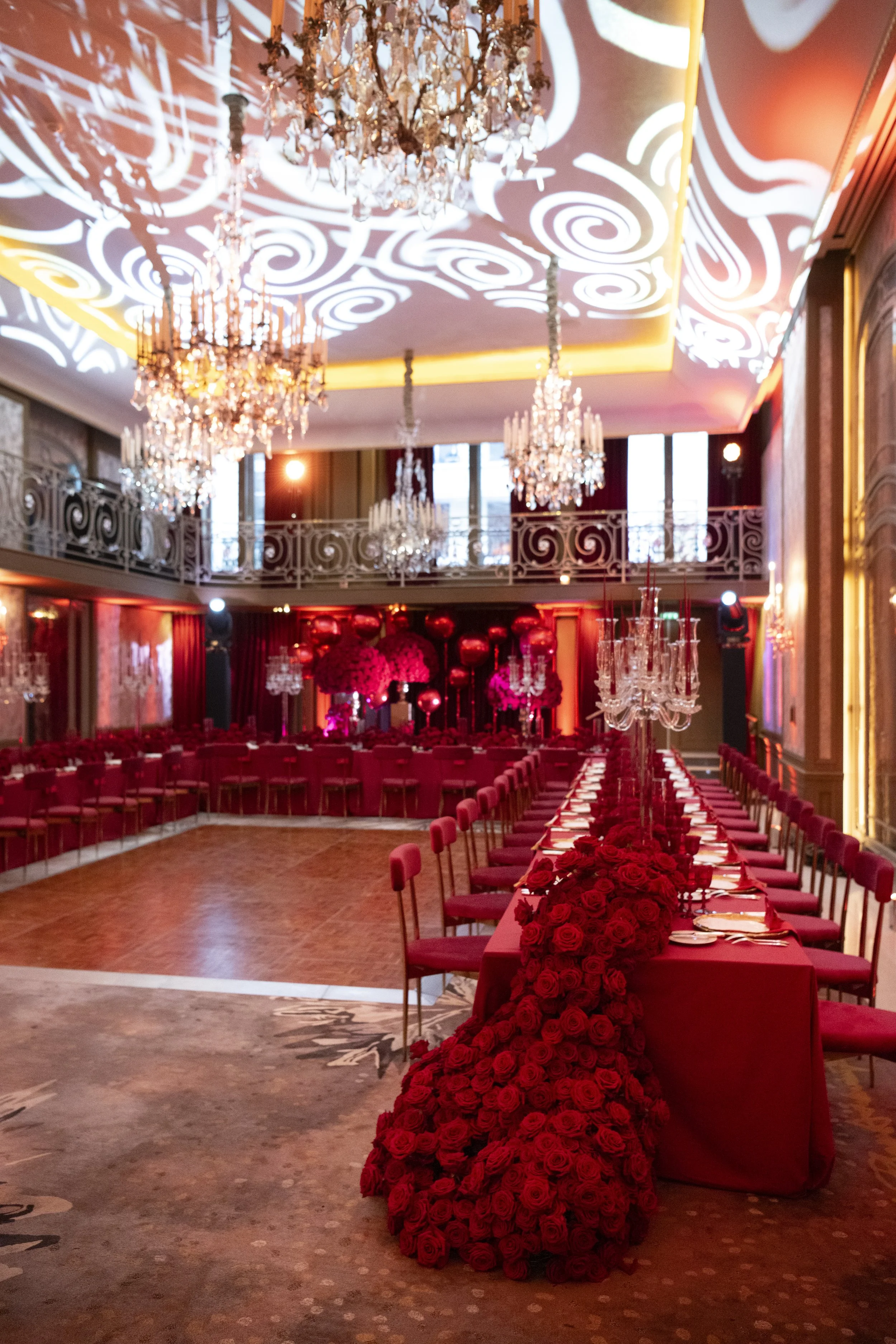 An opulent banquet hall decorated with red roses, chandeliers, and intricate lighting patterns on the ceiling, set up for an elegant event.