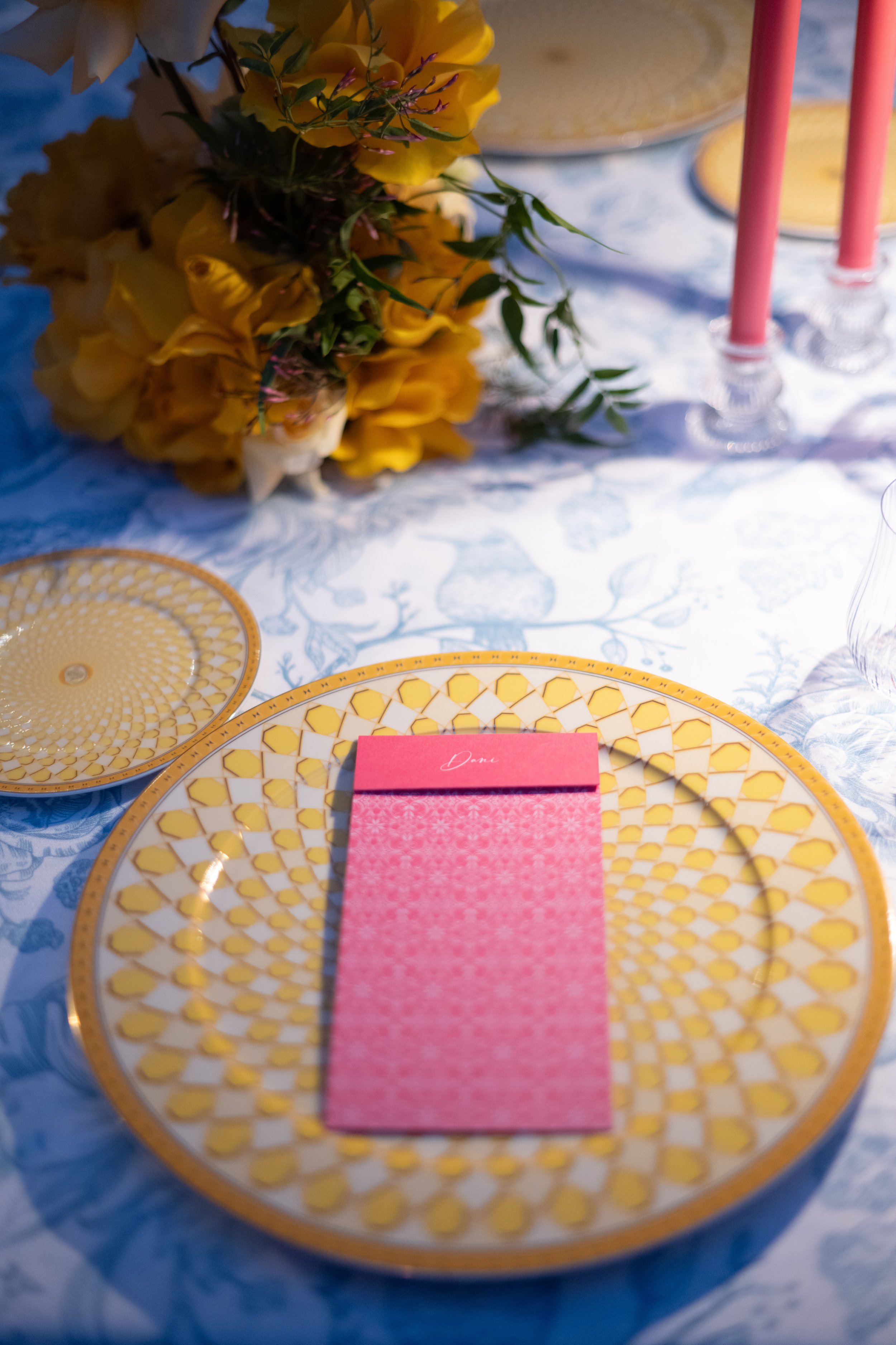 A decorative table setting with a gold and white patterned plate, a pink napkin, and a pink place card with the word 'Dani' written on it, on a tablecloth with a blue floral design. There is a floral arrangement with yellow and white flowers in the b