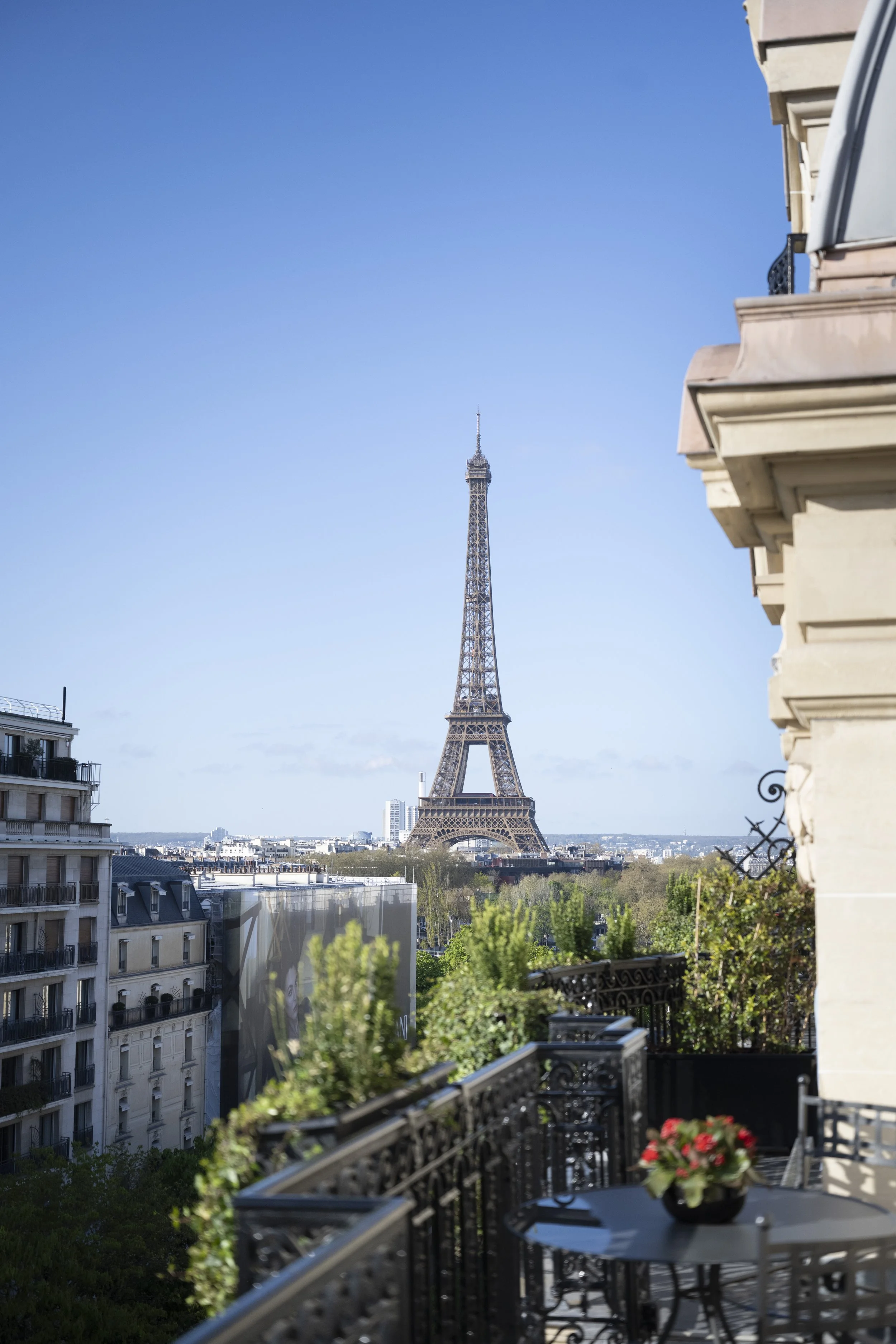 View of the Eiffel Tower from a Paris balcony with plants, a black wrought iron table and chairs, and neighboring buildings under a blue sky.