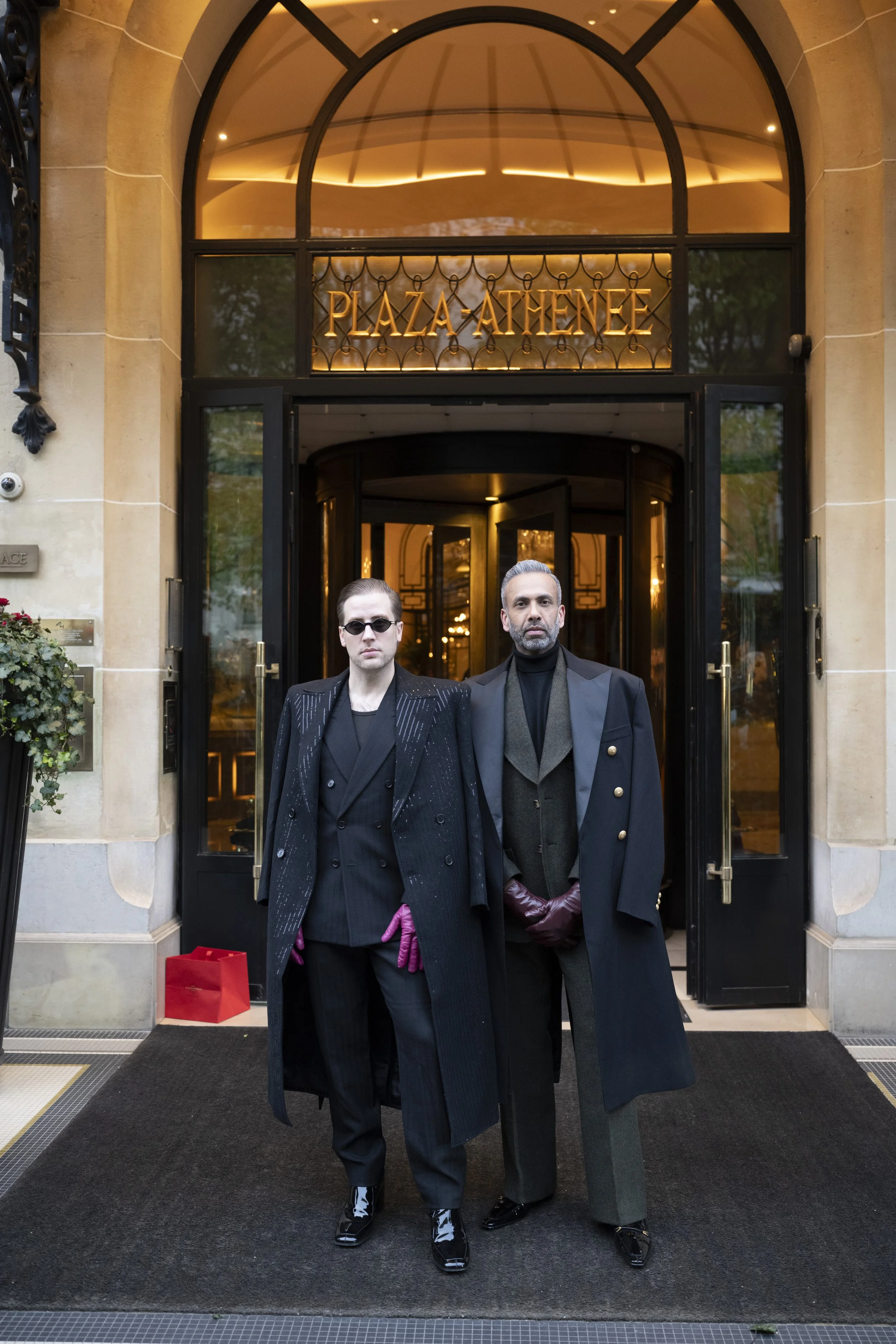 Two men stand in front of the entrance to Plaza Athénée, a luxury hotel. Both are dressed in black suits with stylish coats, and one wears sunglasses with pink gloves. The entrance features large glass doors, ornate lighting inside, and the hotel nam