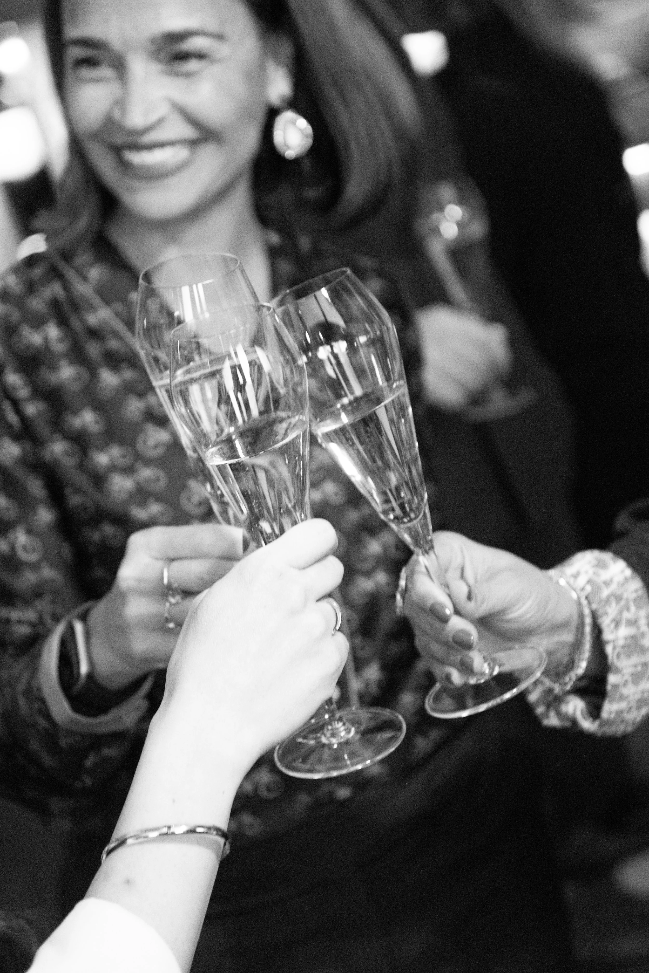 Two women, one with a darker patterned dress and the other with a light top, are clinking glasses of champagne in celebration at a social gathering, with smiling faces and blurred background lights.