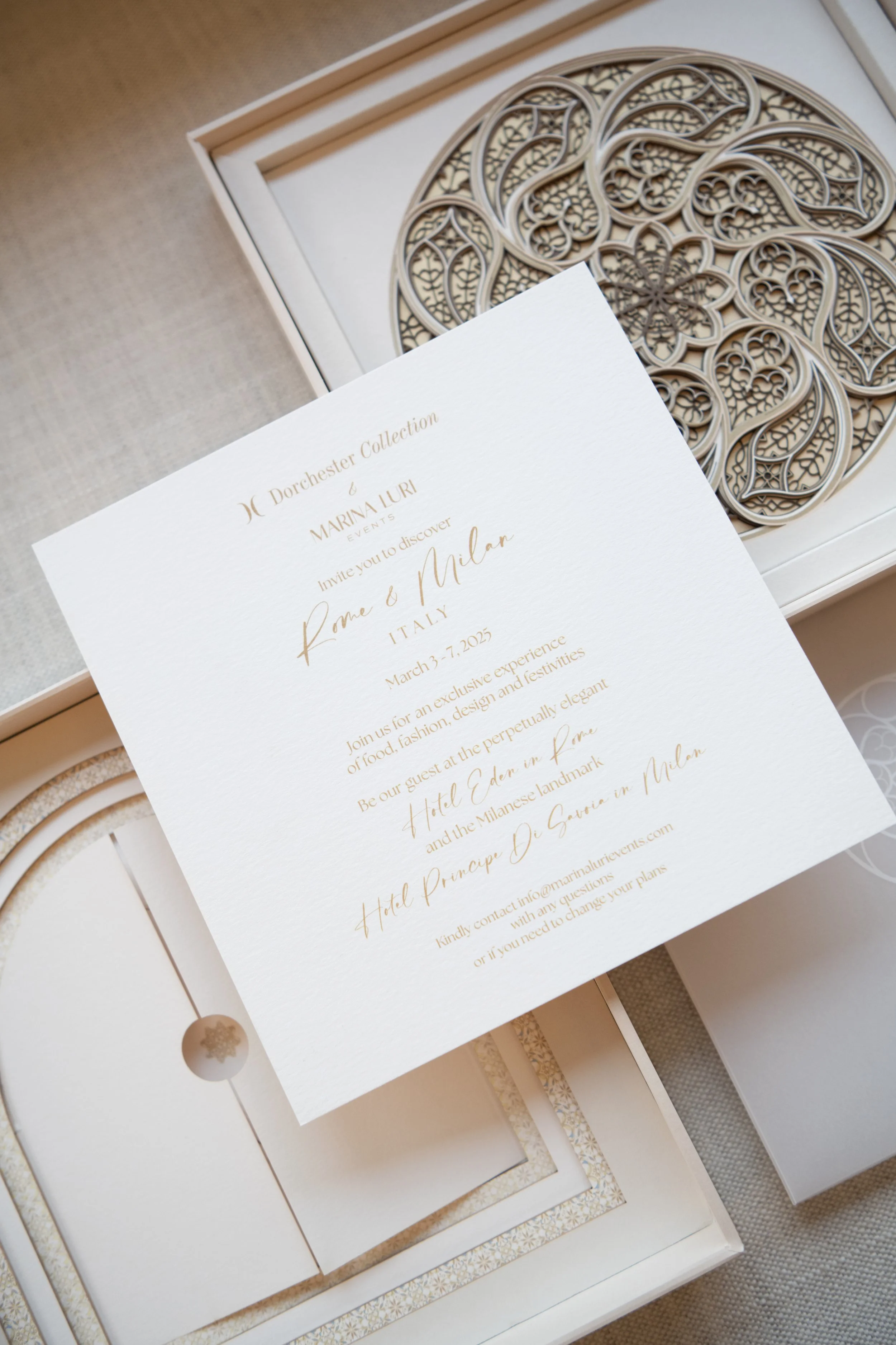 Elegant wedding invitation with gold text, featuring details about an event in Milan, Italy, including date and venue, accompanied by a decorative intricate circular design in the background.