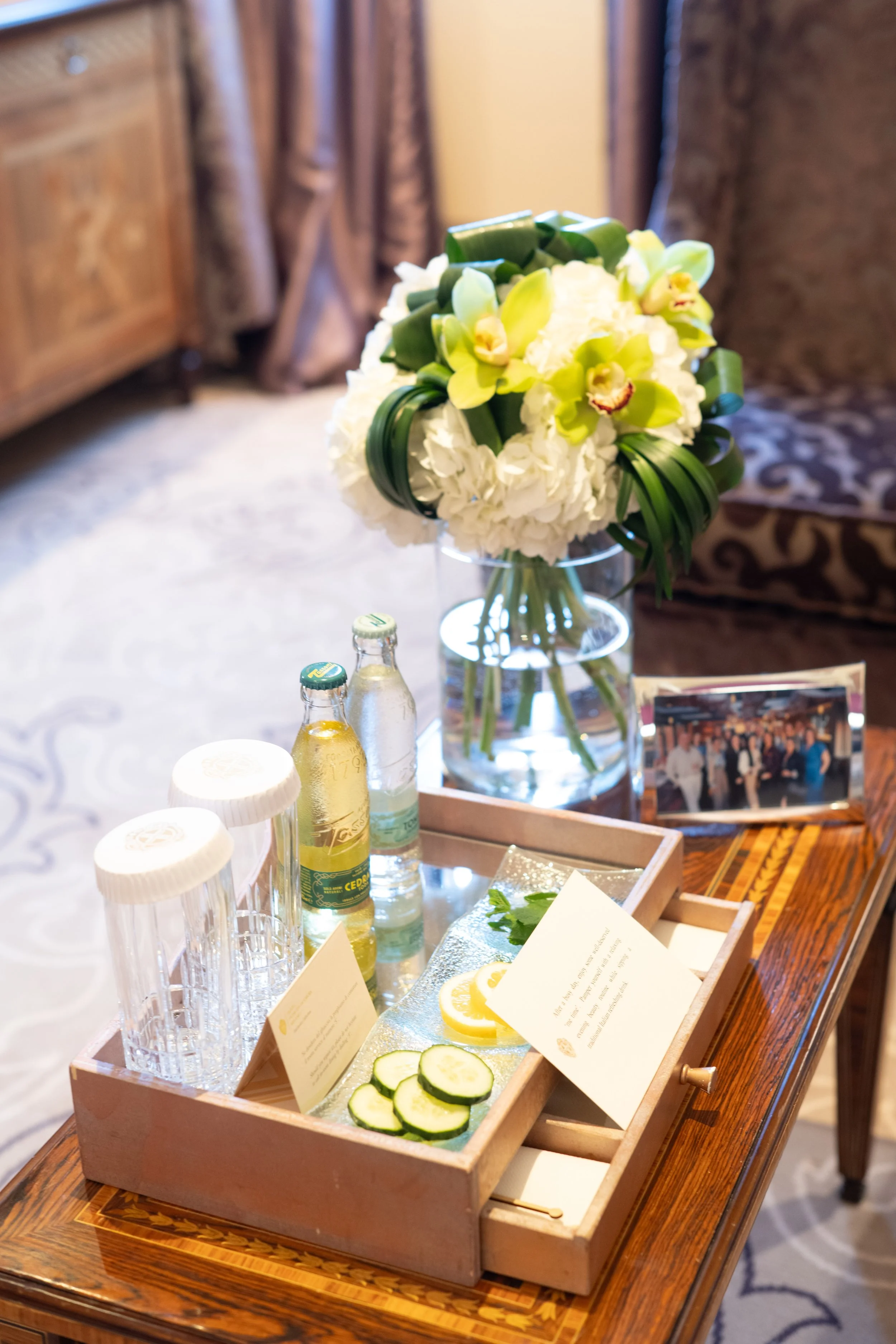 A wooden table holding a pink tray with sliced cucumbers, lemon slices, and informational cards, along with a clear vase of green and white orchids and hydrangeas, silver ice buckets, and bottled beverages, in a room with wood furniture and patterned