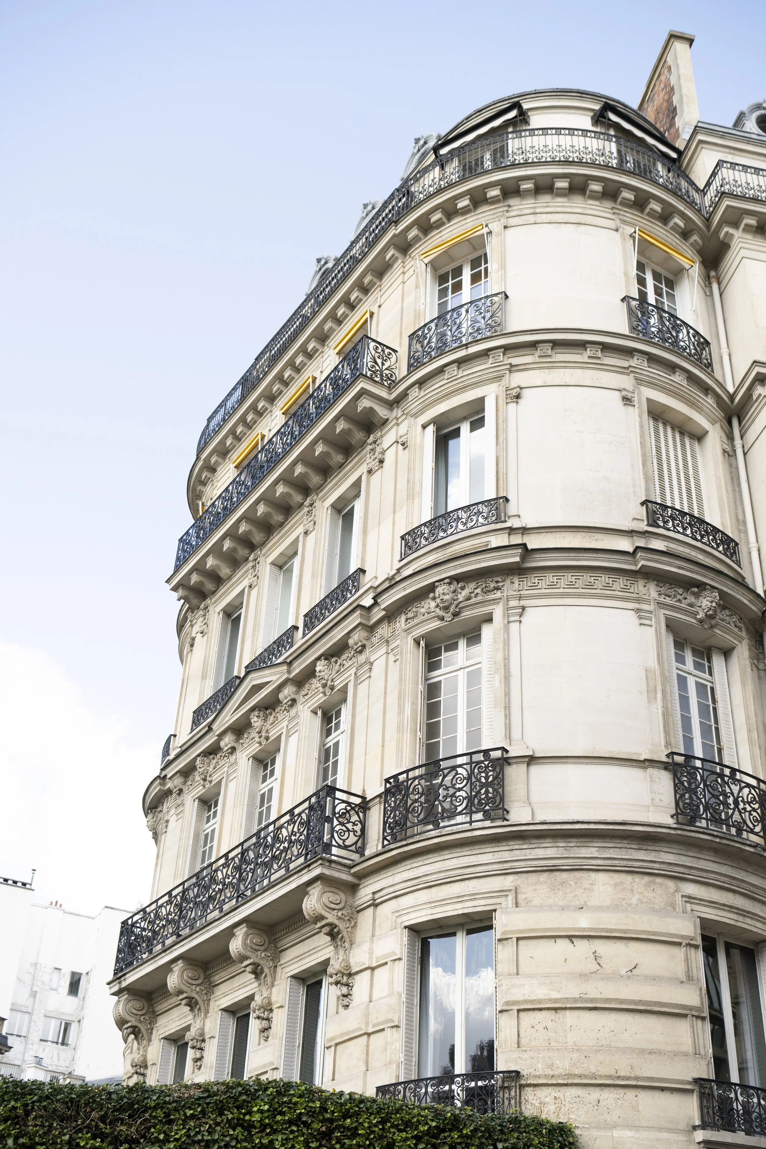 Curved Parisian apartment building with ornate wrought iron balconies and large windows, greenery at the base, under a clear blue sky.
