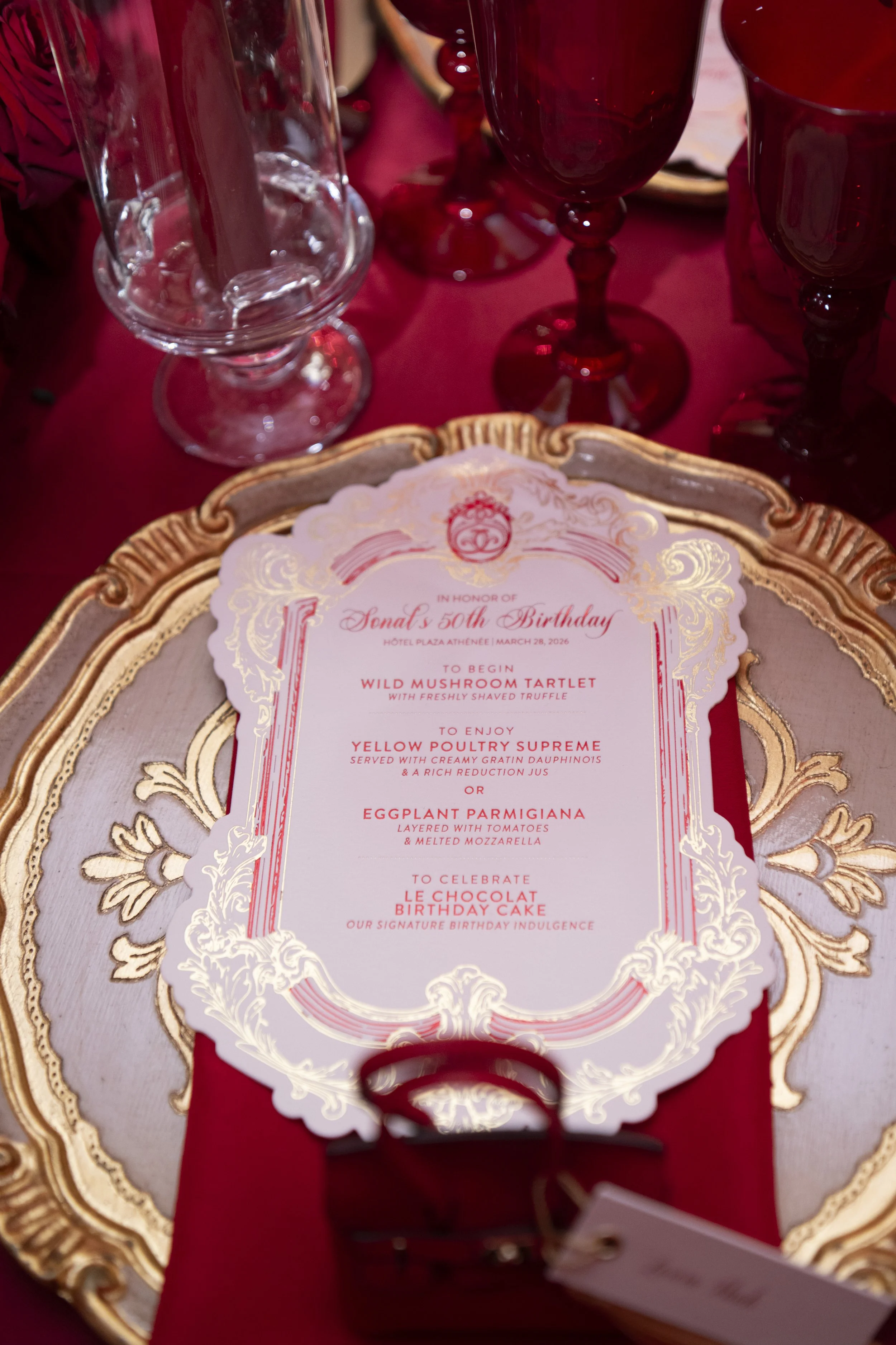 A dining table with ornate gold-trimmed serving tray, red tablecloth, empty clear glass, and dark red goblets, with a birthday menu card for Senat's 50th birthday celebration.