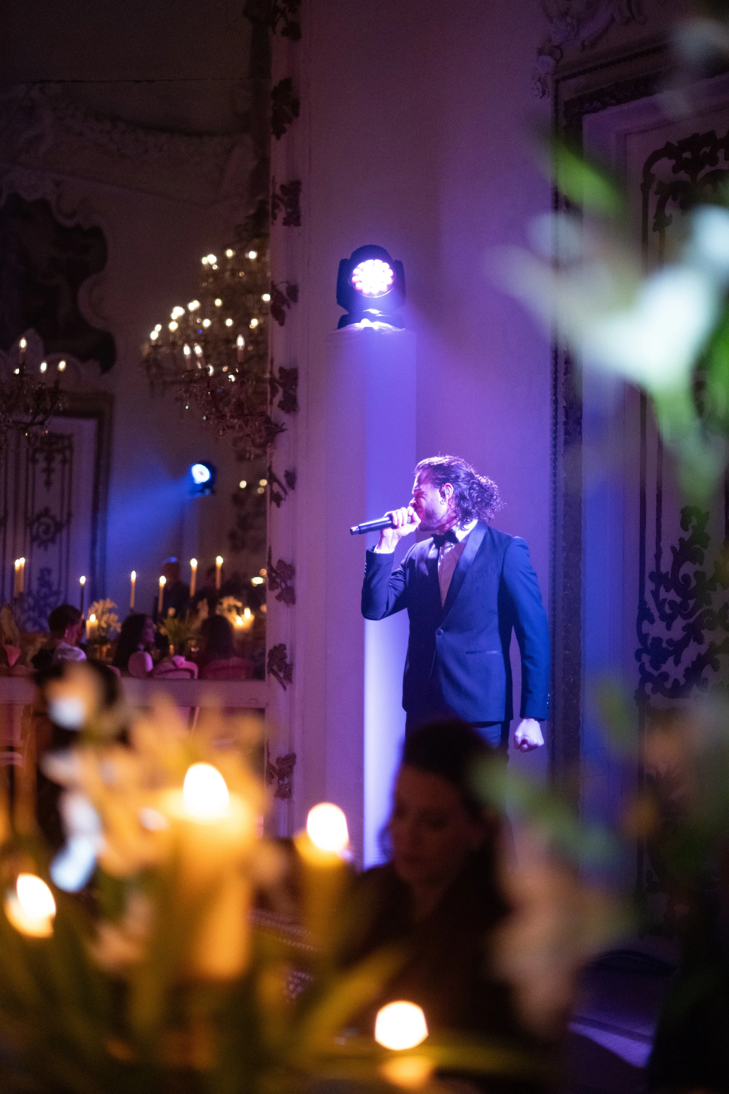 A man in a tuxedo singing into a microphone on stage at an elegant event, with candlelit tables and ornate decor in the background.