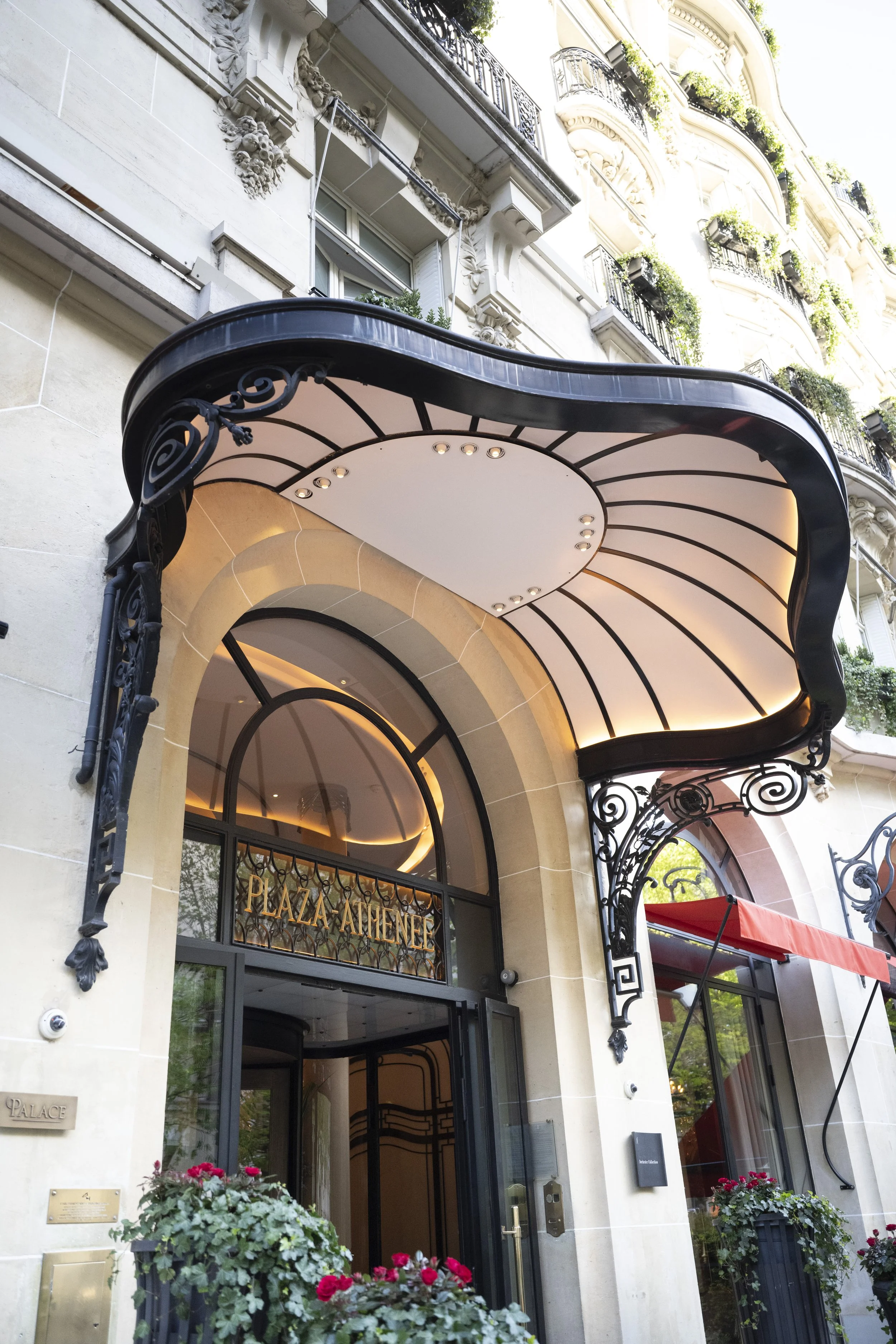 Exterior of a luxurious hotel or building with a black and white awning, ornate wrought iron details, and flower pots with red flowers at the entrance, labeled 'Palace Alleinne' above the doorway.