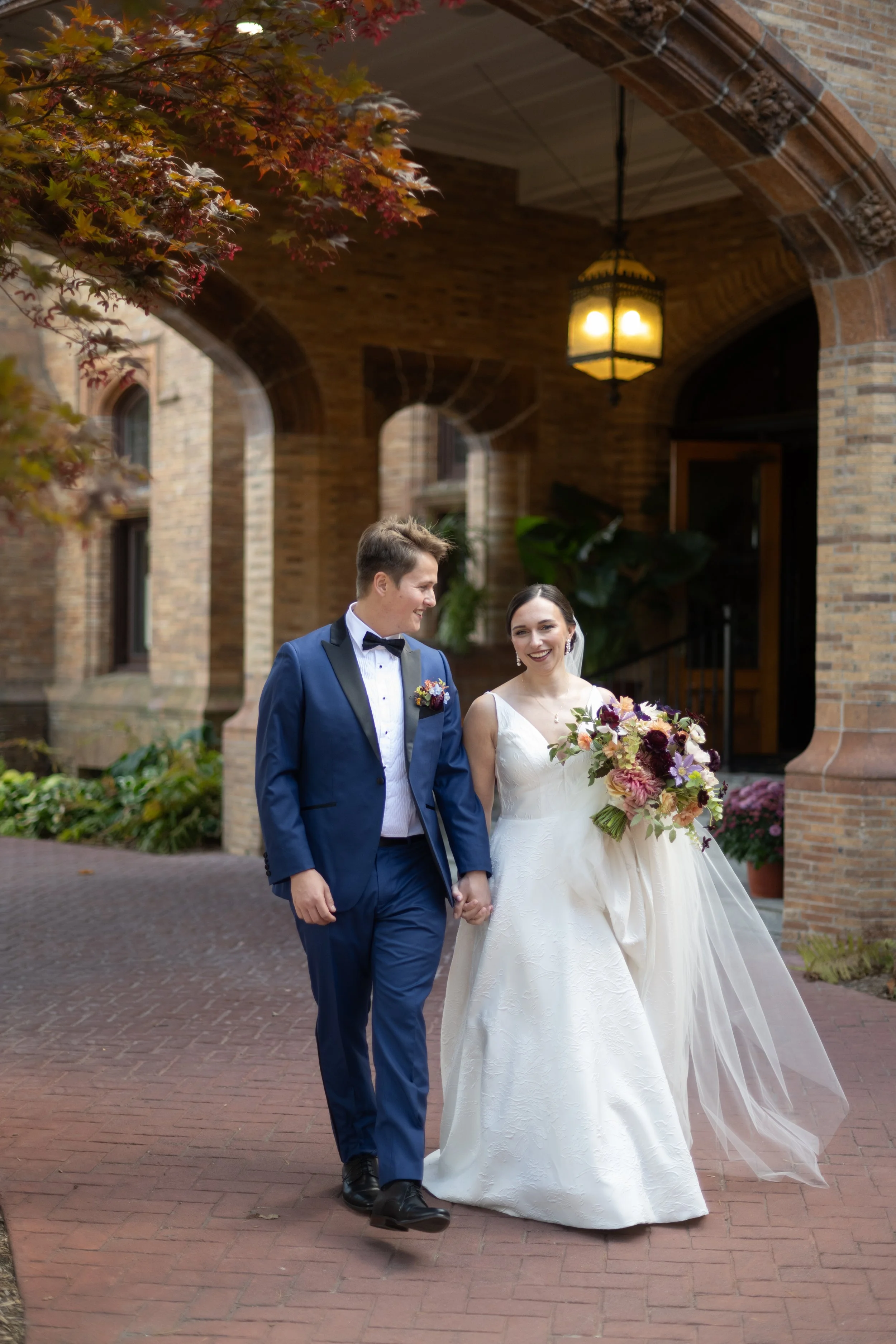 A newlywed couple walking hand in hand outside a brick building during daytime. The groom is wearing a blue suit with a bowtie, and the bride is in a white wedding dress holding a bouquet of flowers. They are smiling at each other under an archway with hanging lanterns and autumn foliage.