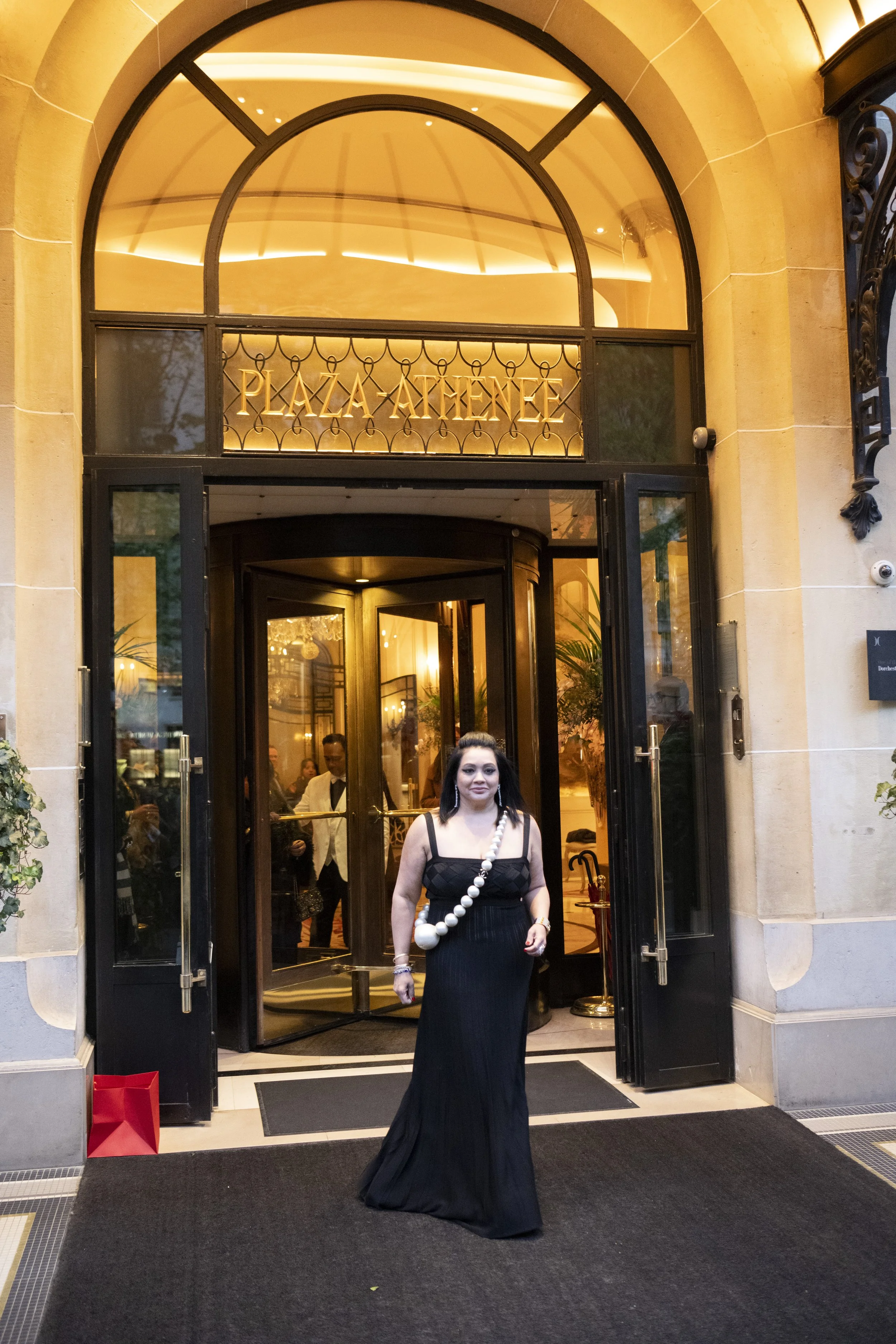 A woman in a black dress with a large bead necklace stands in front of the entrance to a building with a sign that reads 'PLAZA ATHENE'. The entrance has fundraising black doors open, revealing the interior with people inside. There is a red shopping