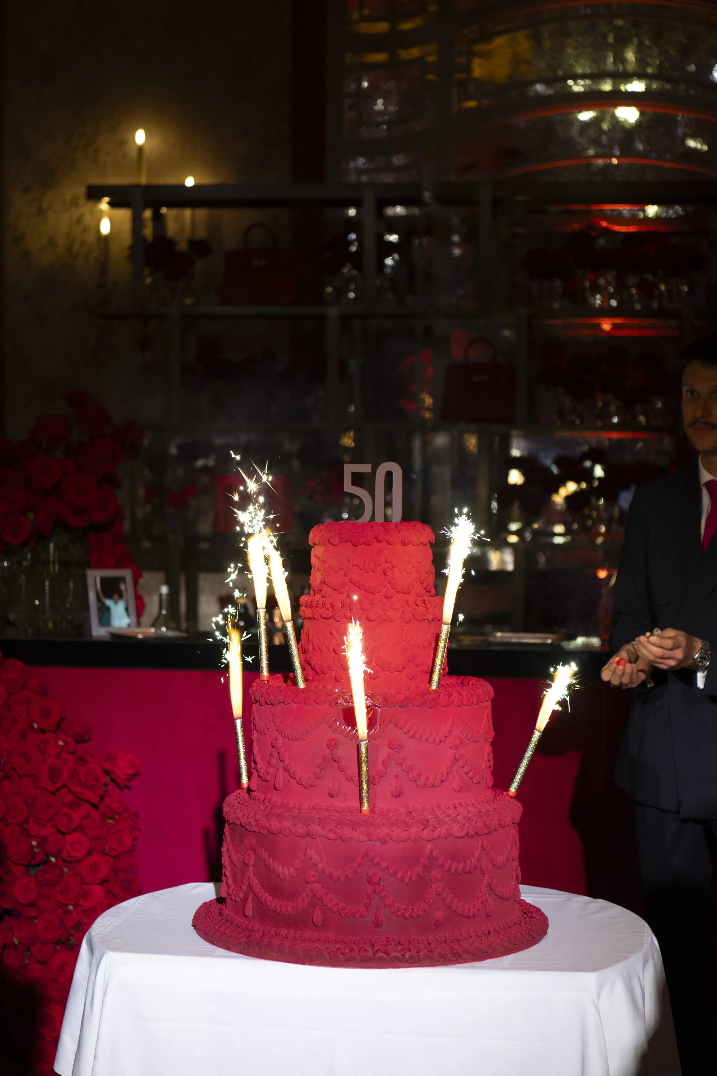 A pink, multi-tiered birthday cake decorated with ruffled icing and a large number 50 topper. Several sparklers are lit on the cake. In the background, there are red roses and a man dressed in a suit with a red tie, celebrating a 50th birthday.