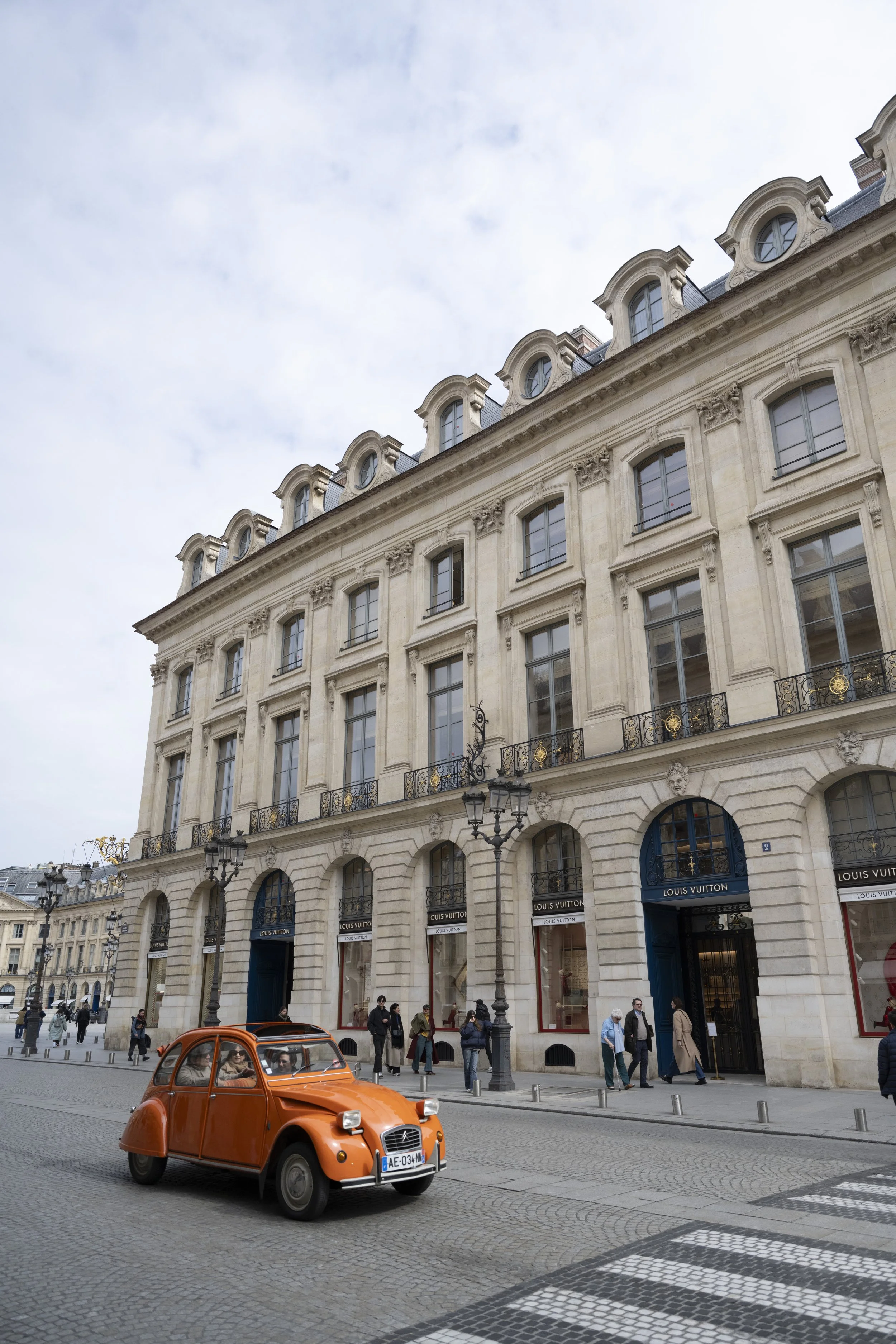 A street scene in front of a Louis Vuitton store in Paris, France, with pedestrians walking and a small orange vintage-style car driving by on cobblestone pavement.