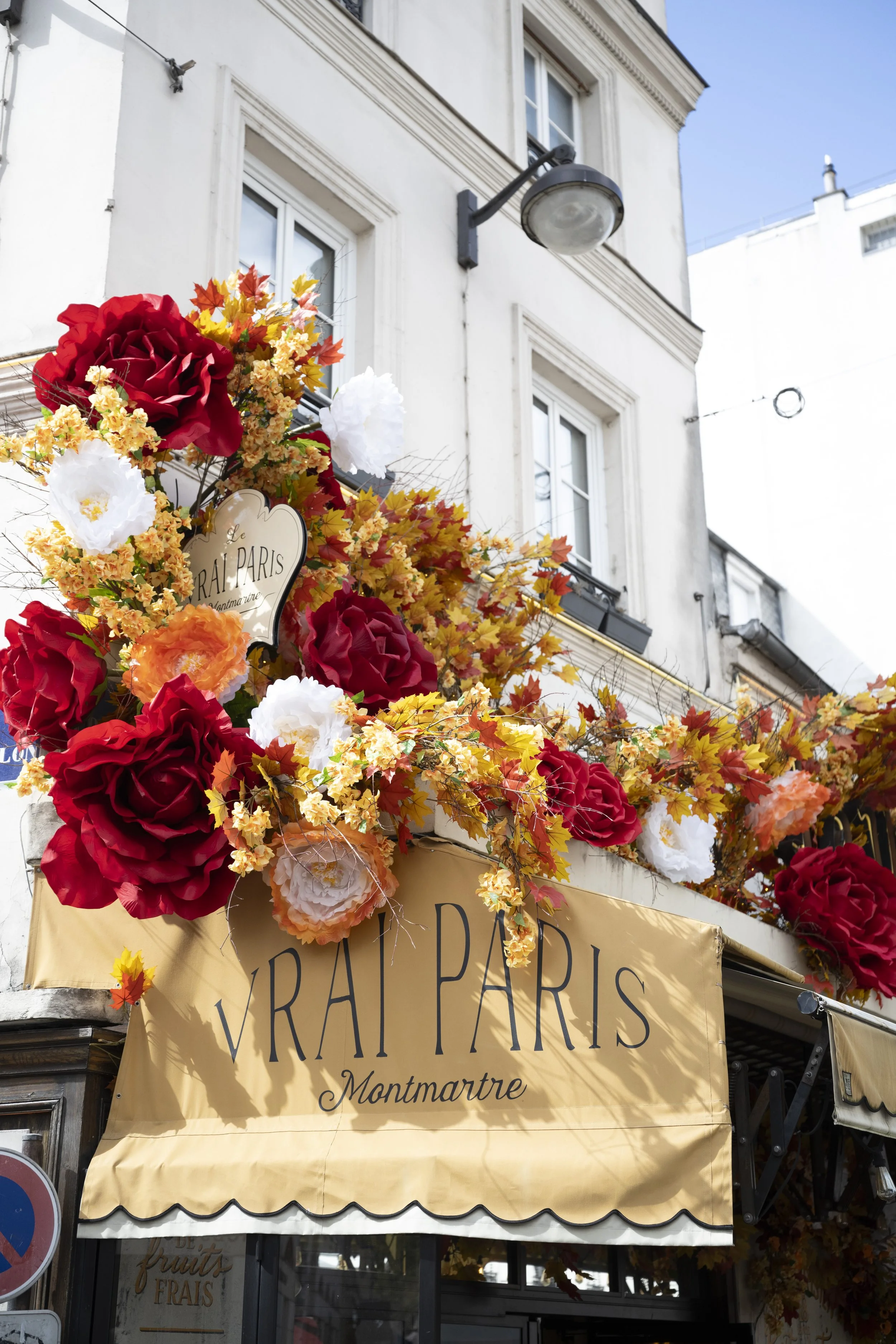 Display of vibrant artificial flowers in shades of red, white, and orange along with autumn leaves, decorating the exterior of a French fruit and vegetable shop in Montmartre, Paris, under a yellow awning with black text.